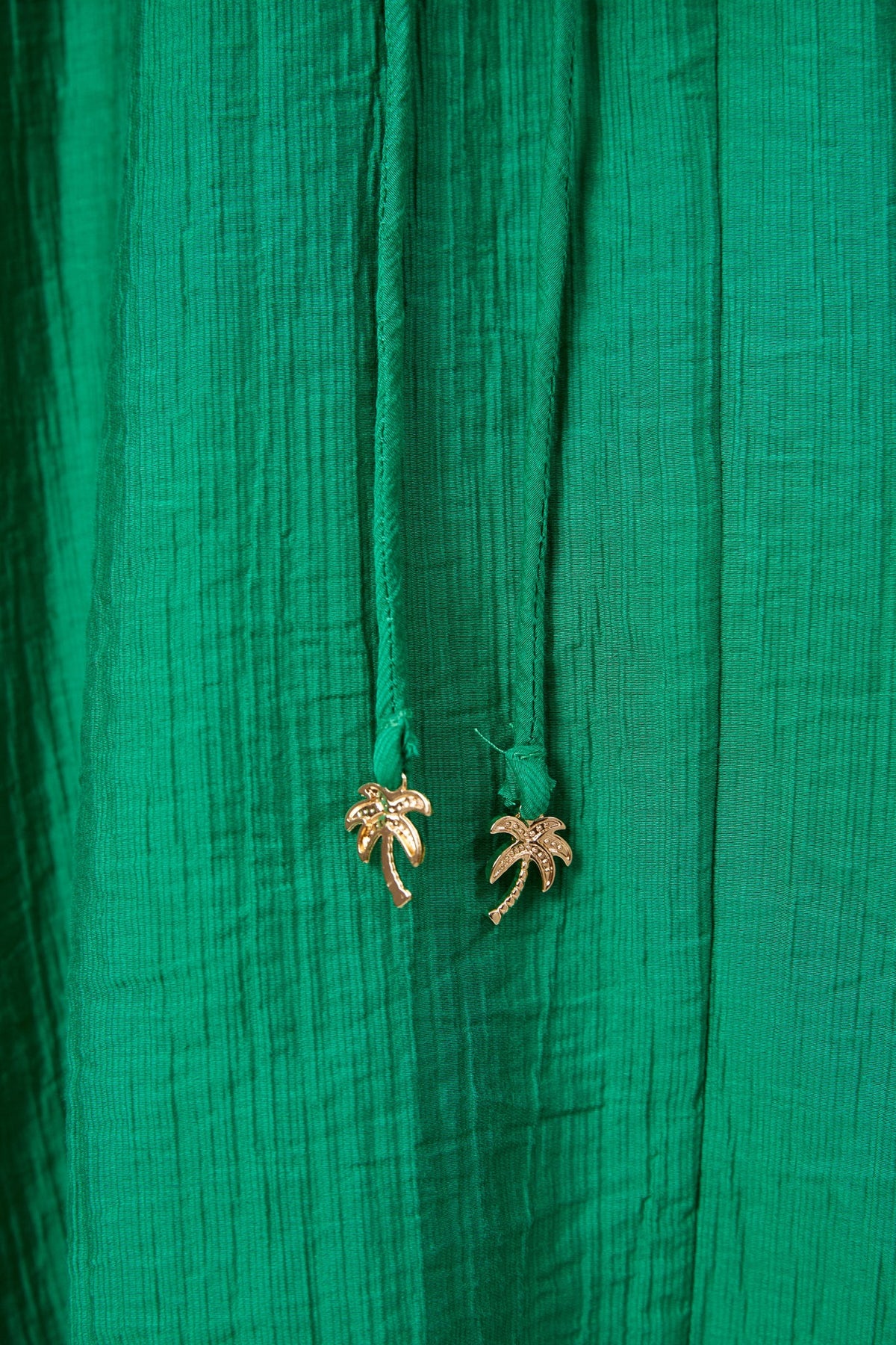 Close-up of a Haven textured green fabric garment with two drawstrings ending in small gold palm tree charms.