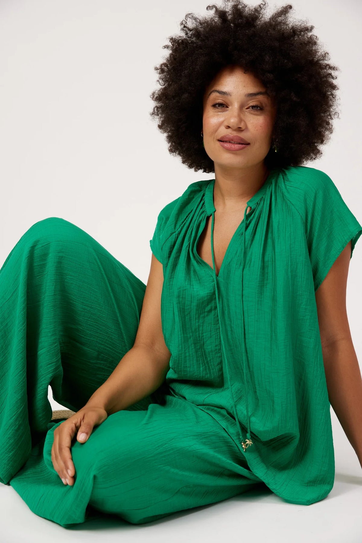 A woman with an afro hairstyle wearing a Haven vibrant green crinkled linen jumpsuit, sitting against a plain white studio background.