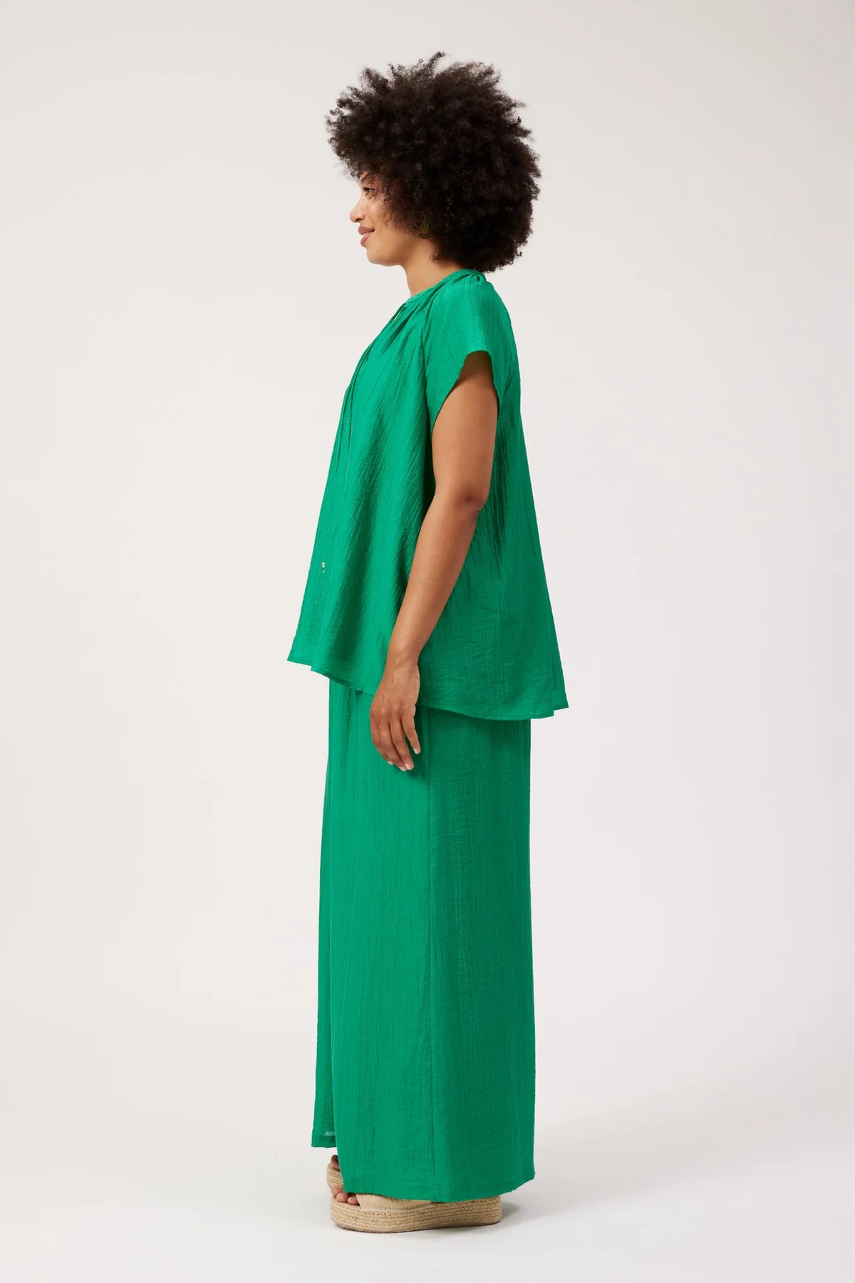 A woman with an afro hairstyle is shown in profile wearing a Haven matching green sleeveless top and wide-leg pants set, standing against a plain light gray studio background.
