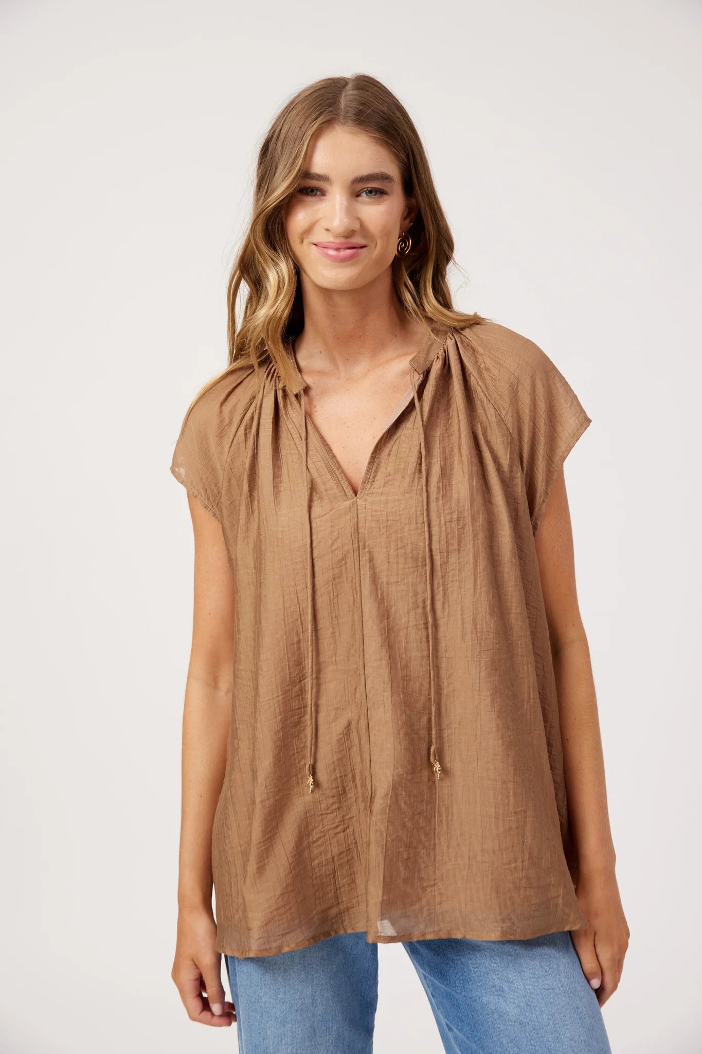 Image of A woman modeling a Haven loose, short-sleeve brown top with a drawstring neckline, paired with blue jeans, against a plain light gray studio background.