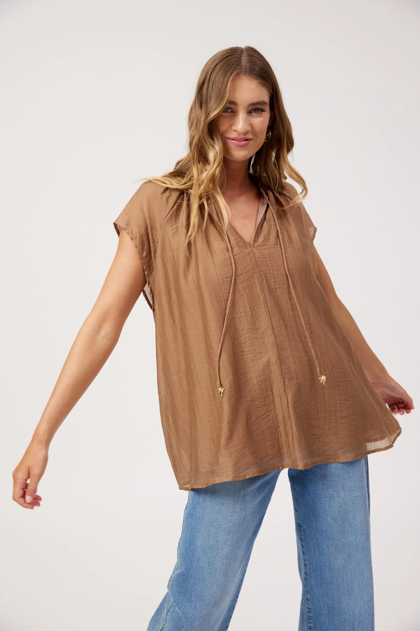 Image of A woman modeling a Haven brown short-sleeve V-neck top with drawstrings, paired with blue jeans, against a plain light gray studio background.