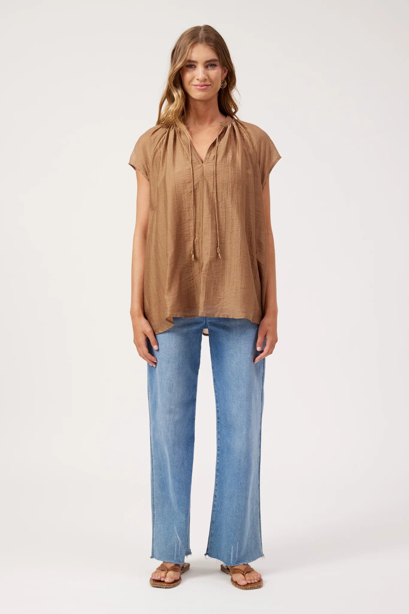 Image of A woman modeling a Haven loose, short-sleeve brown blouse with a tie-neck detail, paired with wide-leg blue jeans and brown sandals, against a plain white studio background.