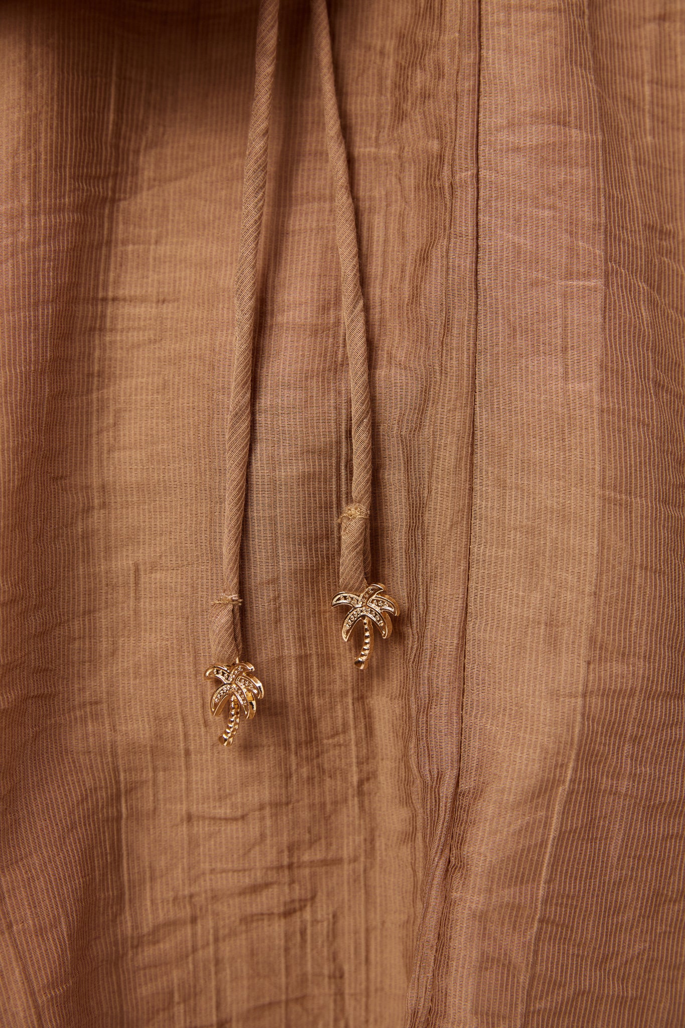 Image of Close-up of a Haven brown textured fabric garment with a drawstring and small gold palm tree charms at the ends.