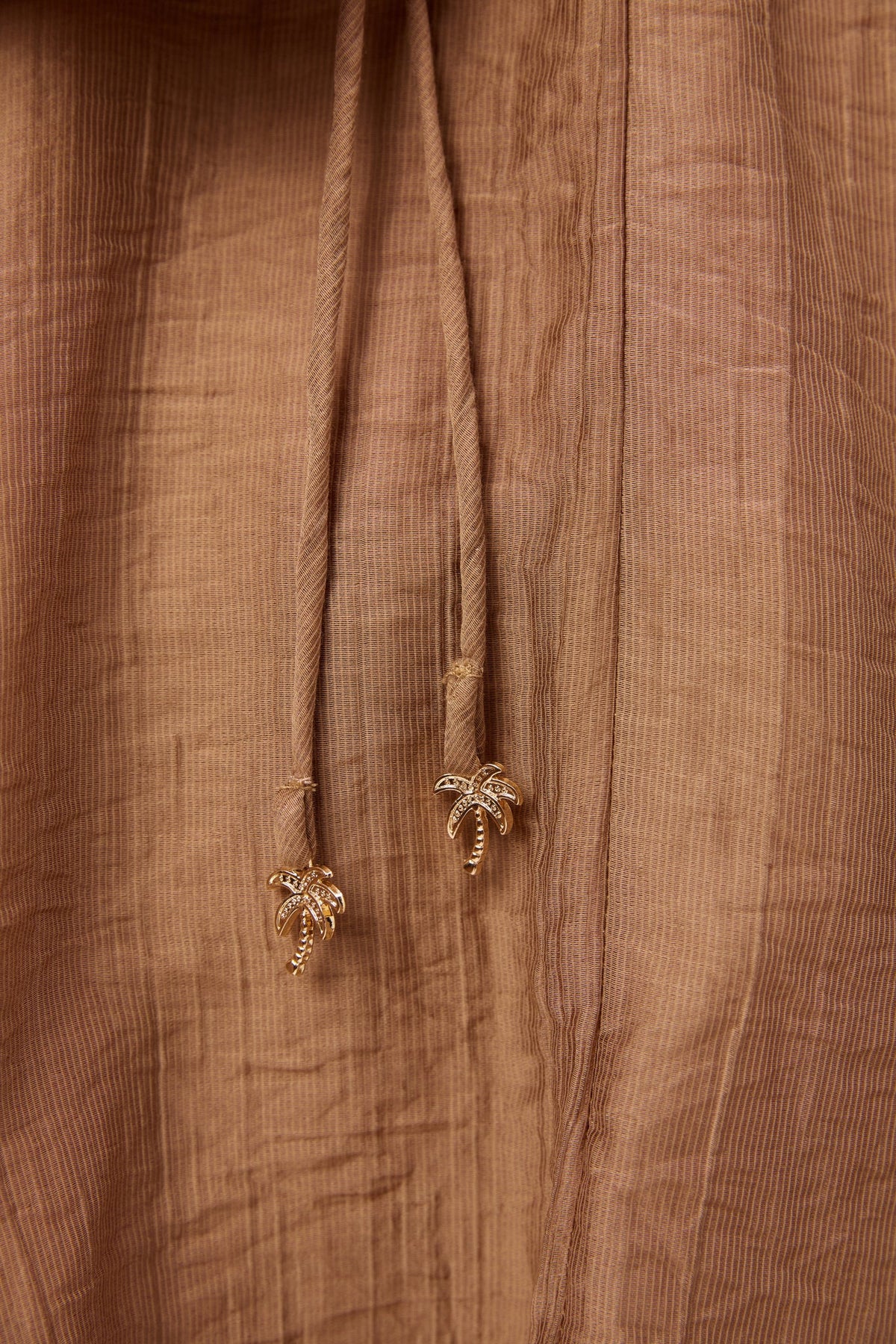 Close-up of a Haven brown textured fabric garment with a drawstring and small gold palm tree charms at the ends.
