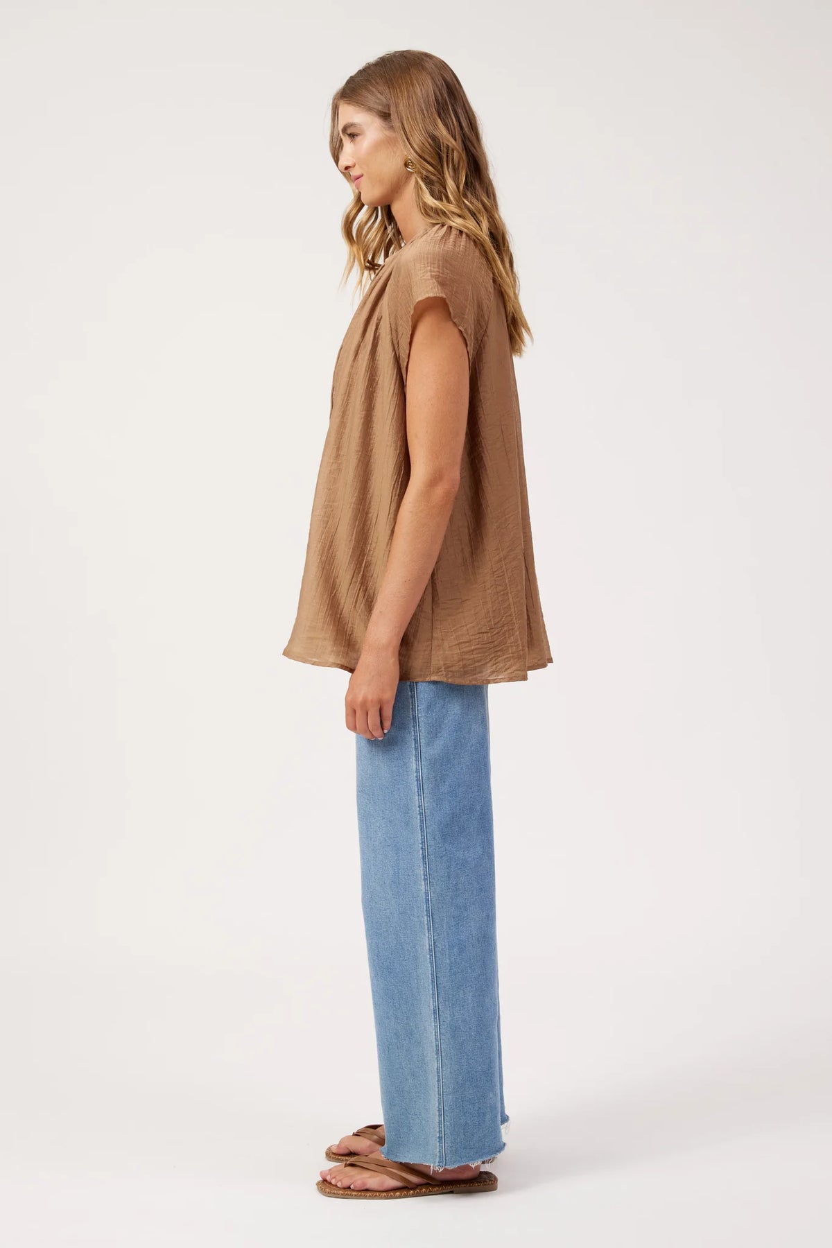 A woman stands in profile wearing a Haven loose, short-sleeve brown top, wide-leg light blue jeans, and brown sandals against a plain white studio background.