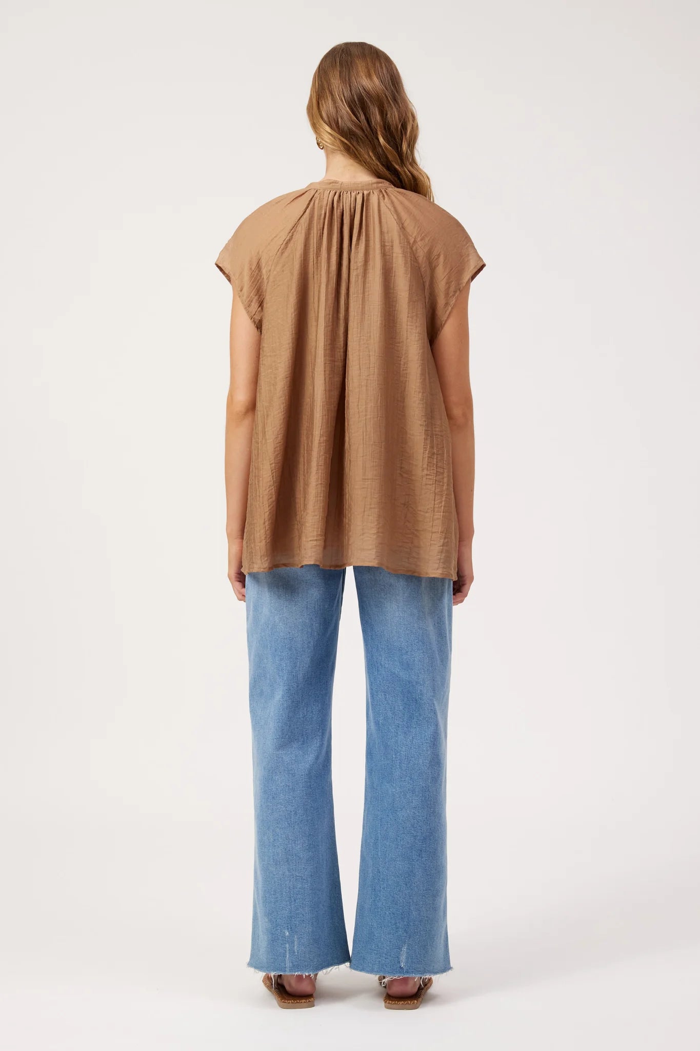 Image of A woman shown from the back wearing a Haven brown sleeveless top with a gathered neckline and wide-leg blue jeans, standing against a plain light background.