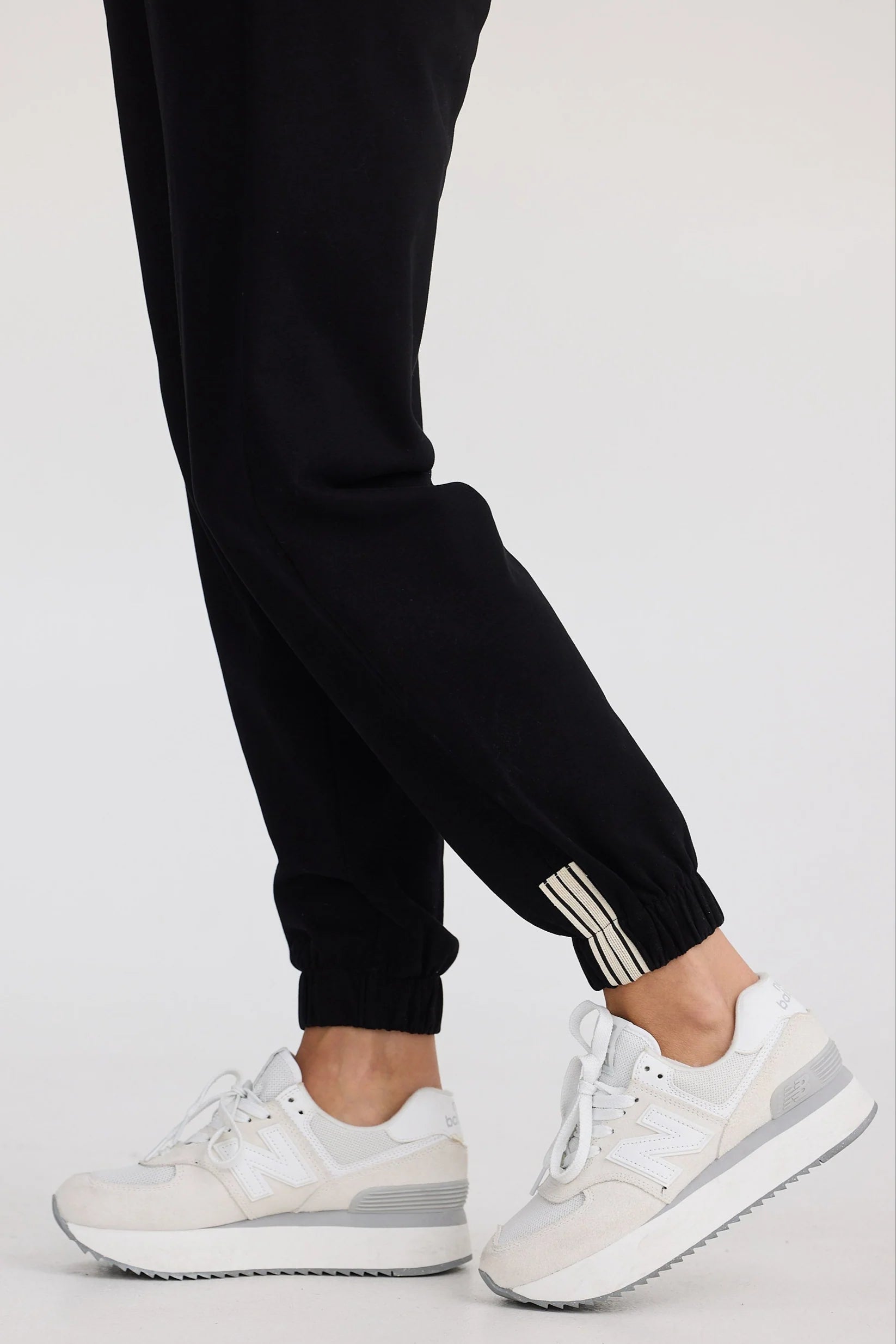 Person wearing black jogger pants with a stripe and white sneakers on a light gray background