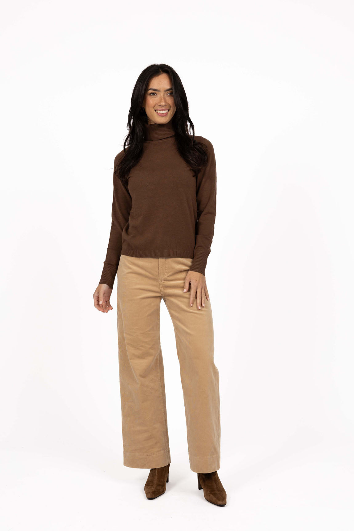 Woman wearing a brown turtleneck and Humidity beige pants on a white background
