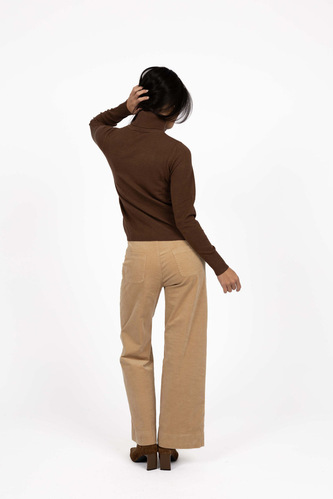 Image of Woman wearing a brown turtleneck and Humidity beige pants on a white background