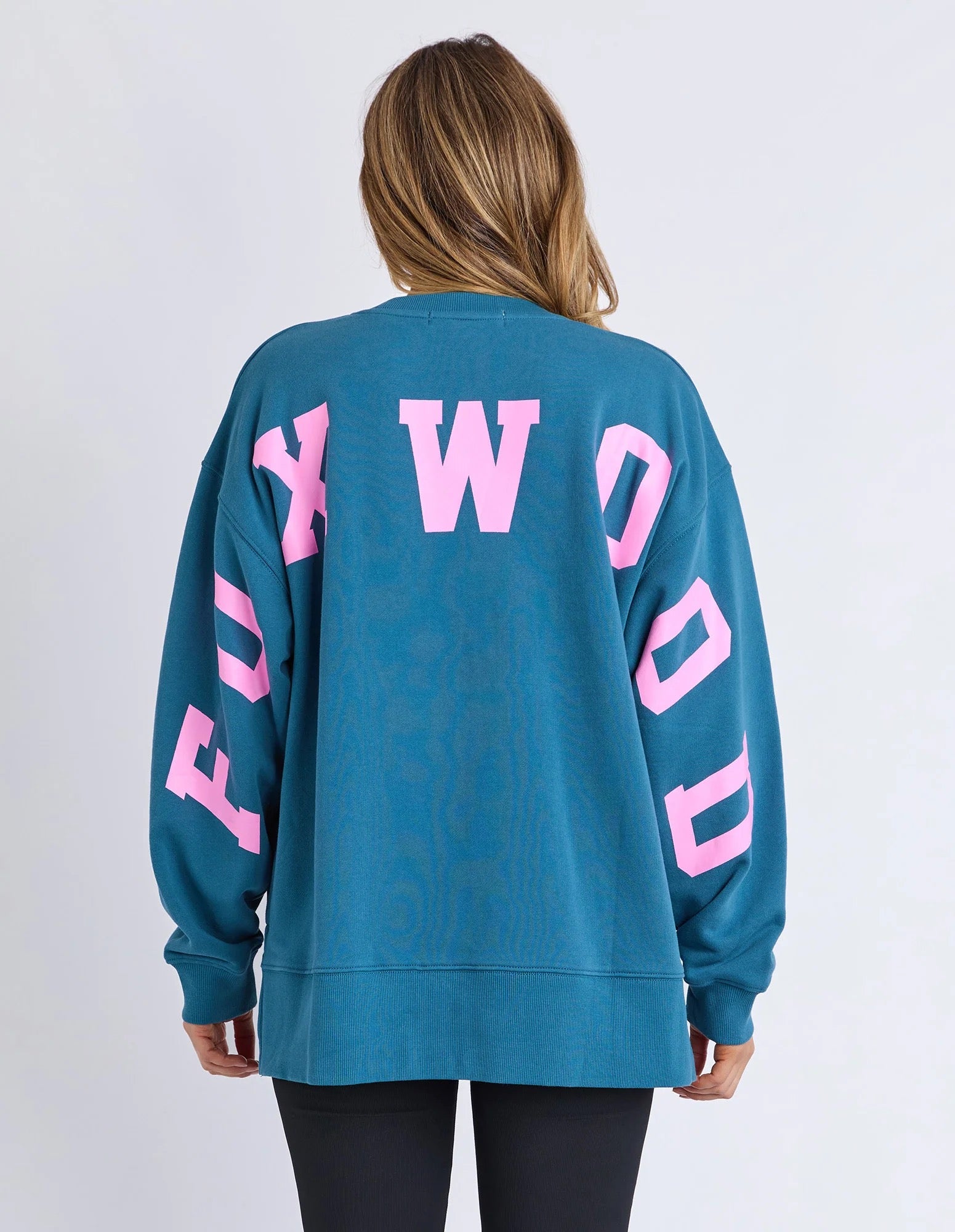 Woman wearing a blue jumper with pink text on the back against a light grey background