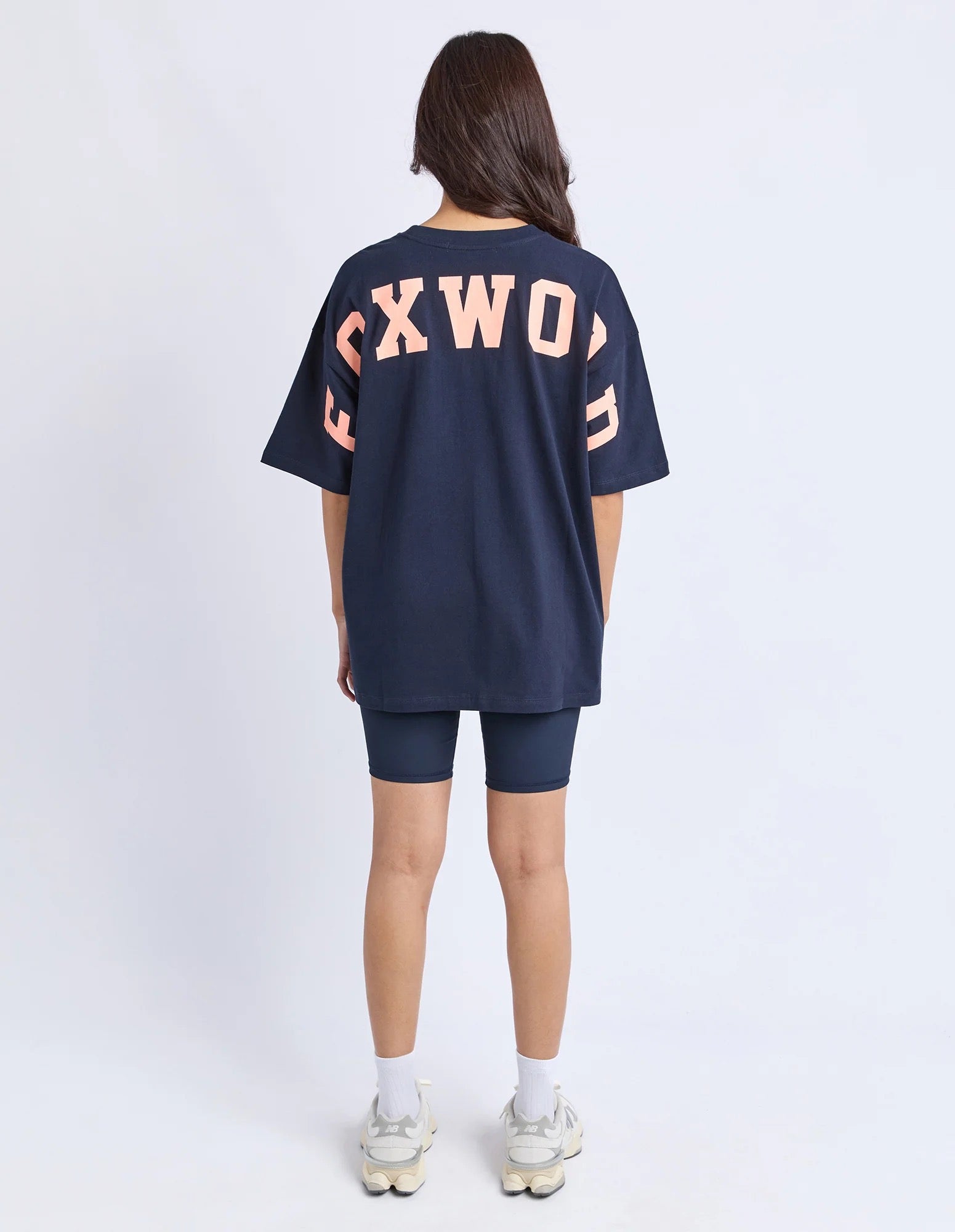 Back view of a woman wearing a navy blue oversized t-shirt with 'FOXWOOD' printed on the backin peach, standing against a white background.