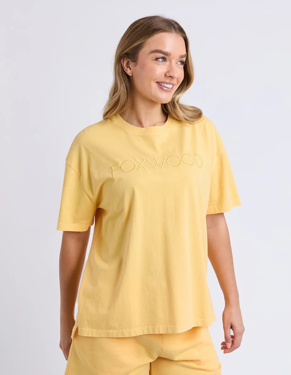 Image of A woman wearing a yellow short-sleeve t-shirt with 'FOXWOOD' printed across the chest in a matching light tone against a light grey studio background.