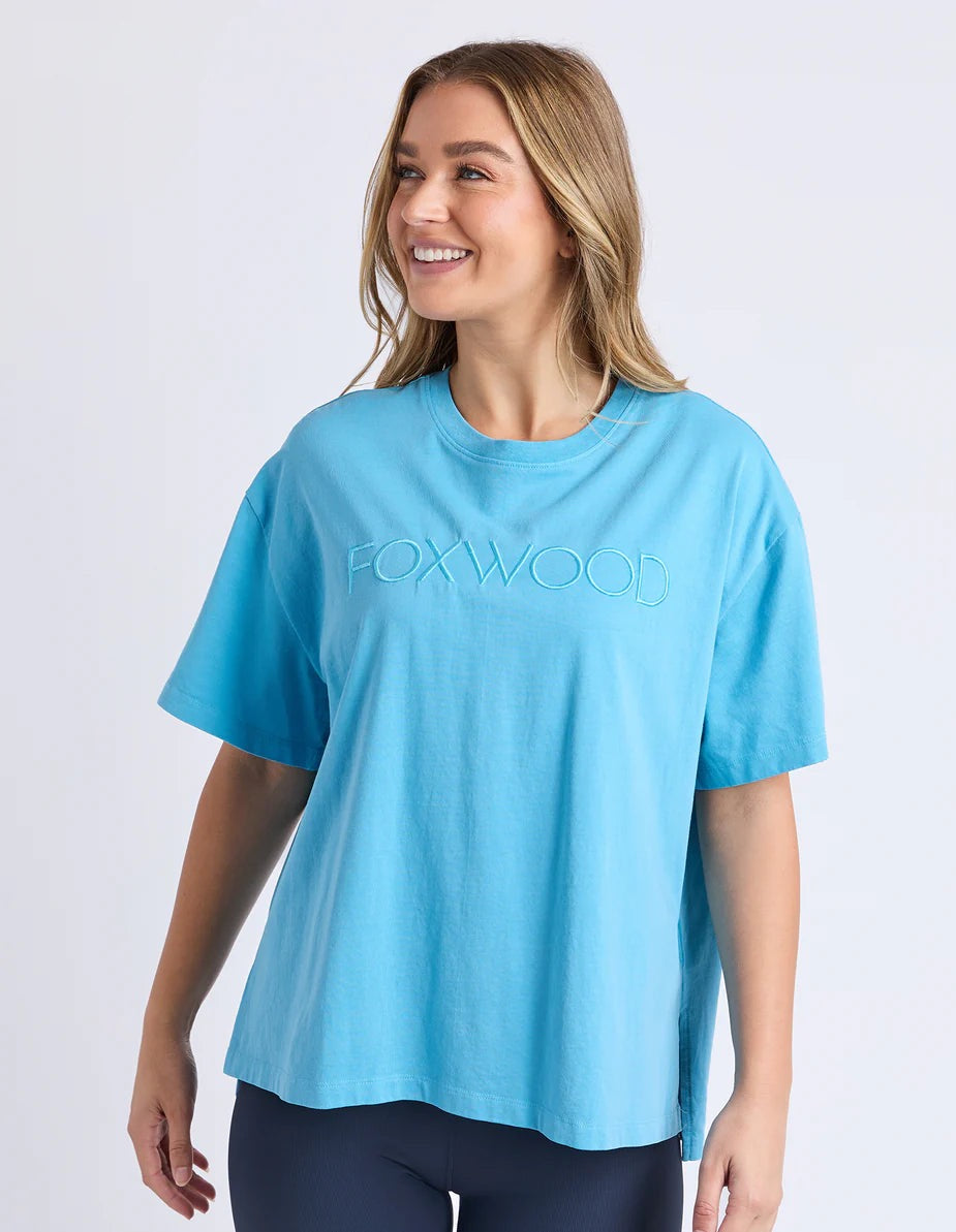 A woman wearing a bright blue short-sleeve t-shirt with 'FOXWOOD' embossed across the chest against a light grey background.