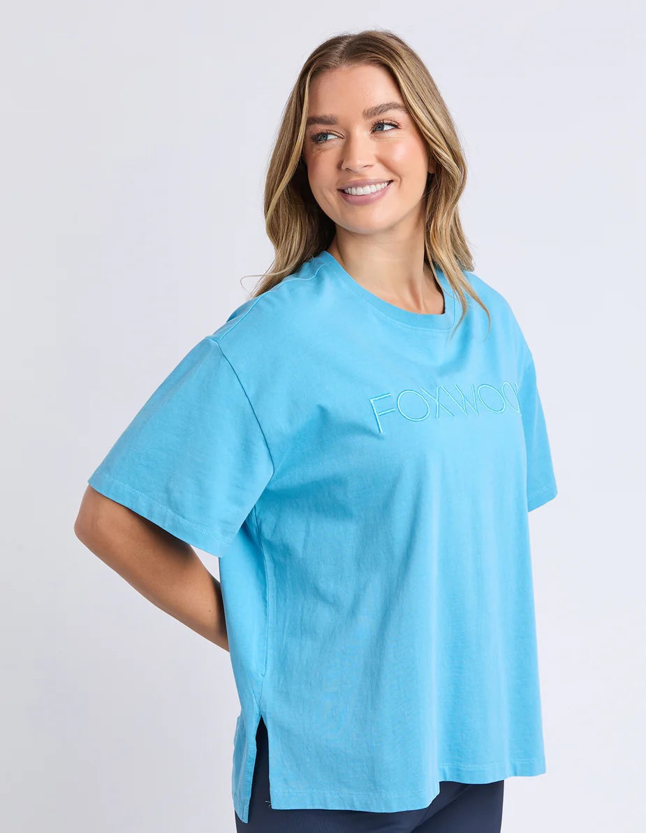 Image of A woman wearing a bright blue short-sleeve t-shirt with a tonal embroidered 'FOXWOOD' logo on the chest, posed against a light grey studio background.