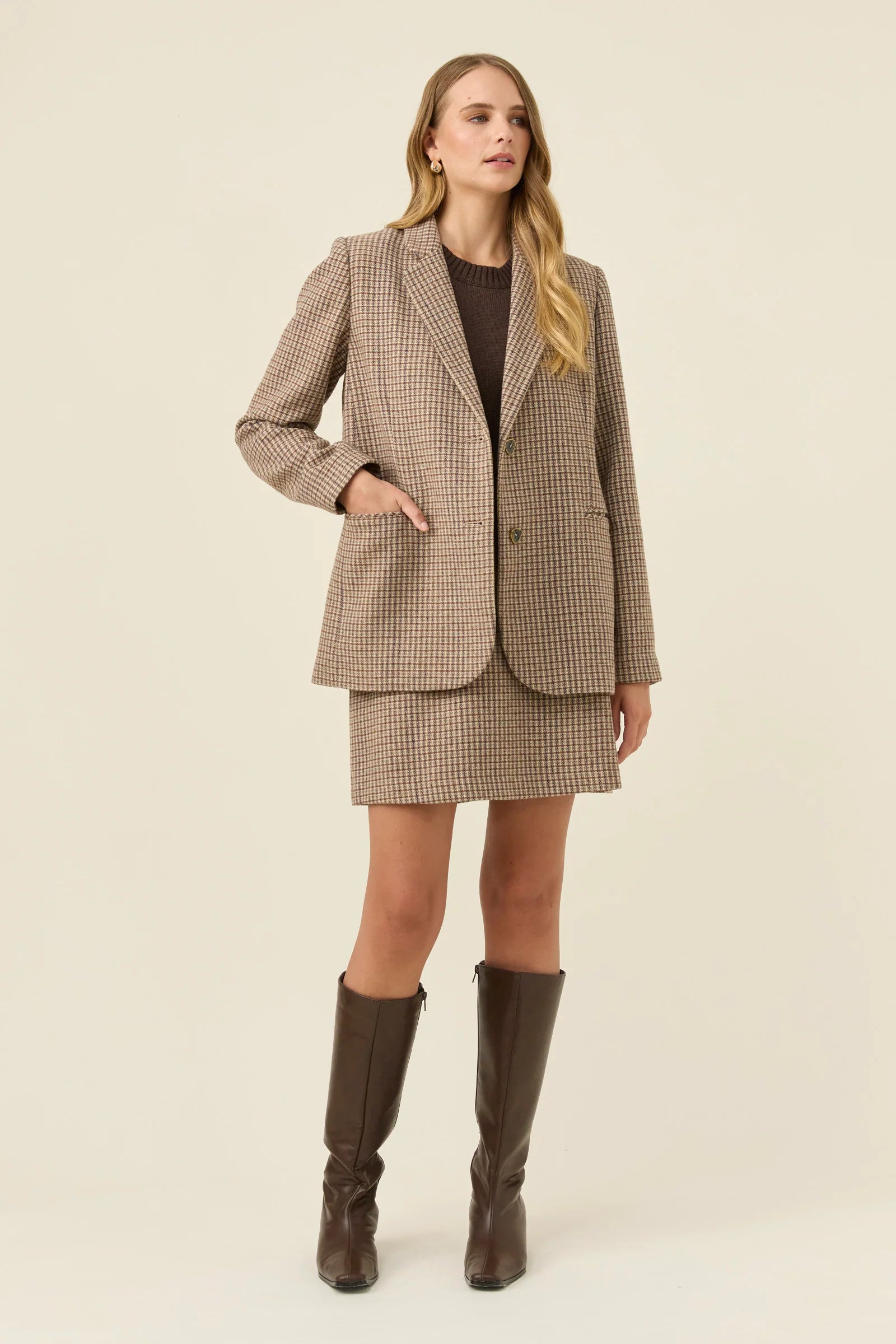 Image of Woman wearing a Isle of Mine brown checkered suit with knee-high boots on a beige background