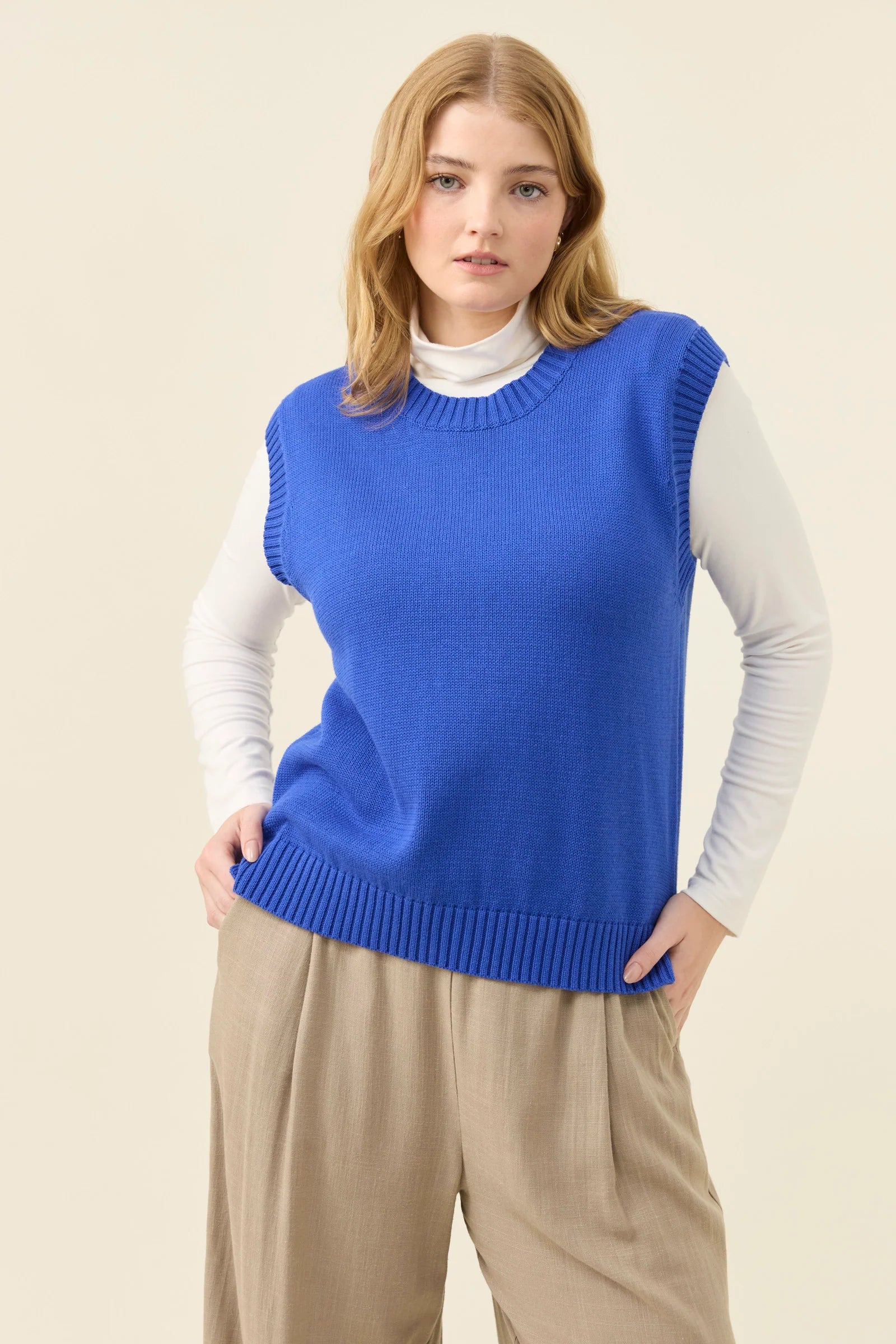 Woman wearing a Isle of Mine blue sweater vest over a white shirt with beige pants on a beige background