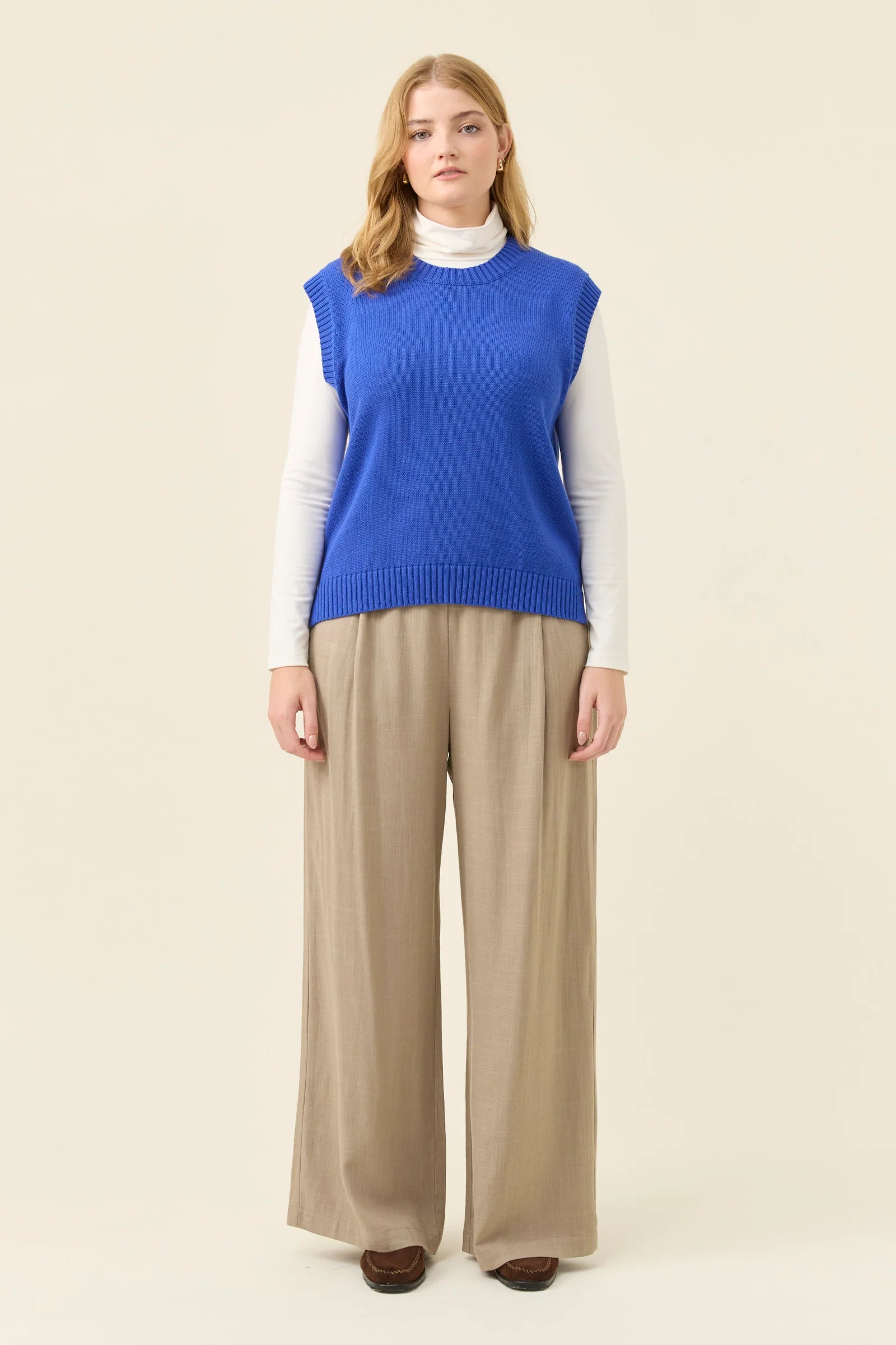 Woman wearing a Isle of Mine blue sleeveless sweater over a white shirt with beige pants on a beige background