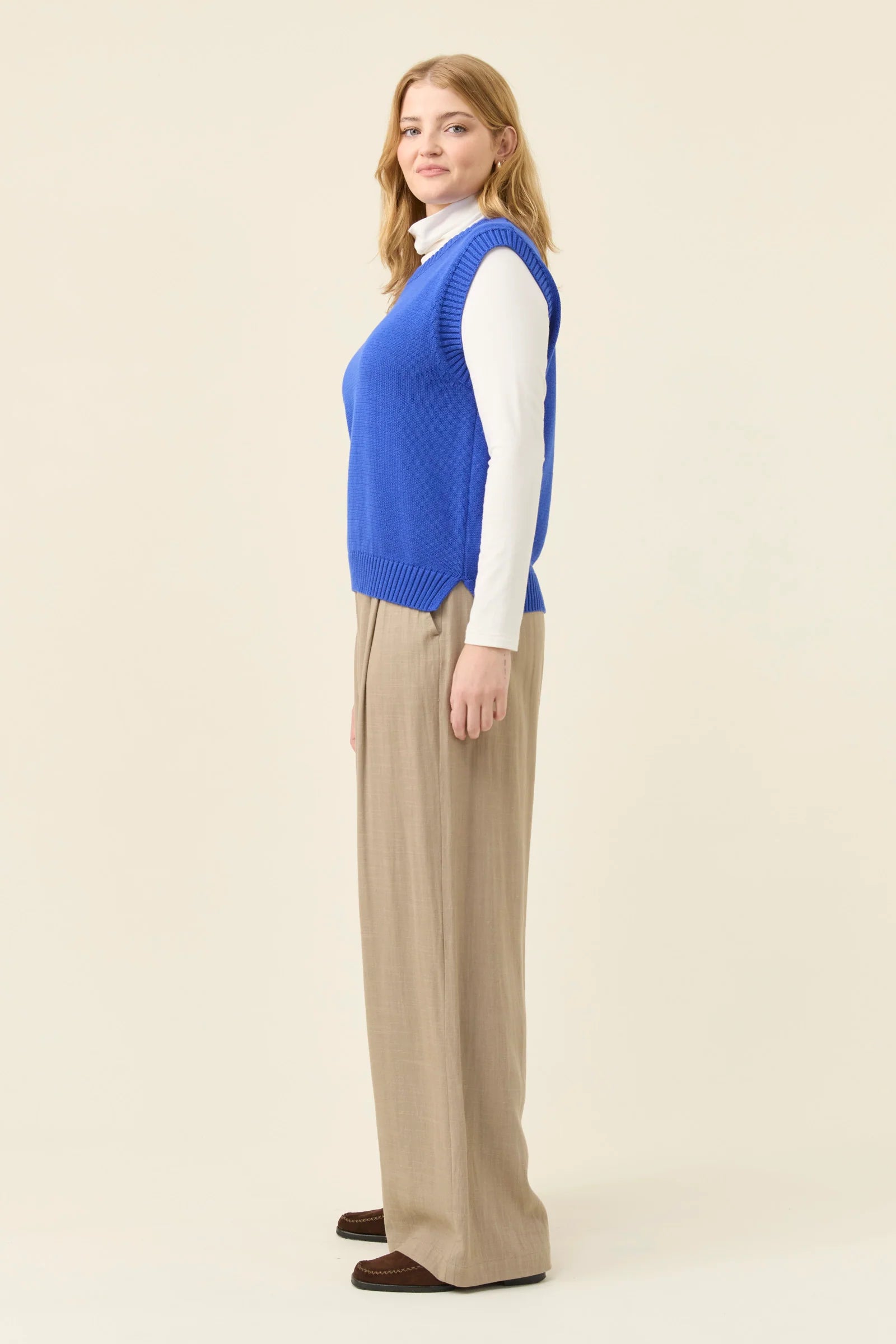 Woman wearing a Isle of Mine blue vest over a white shirt with beige pants on a beige background