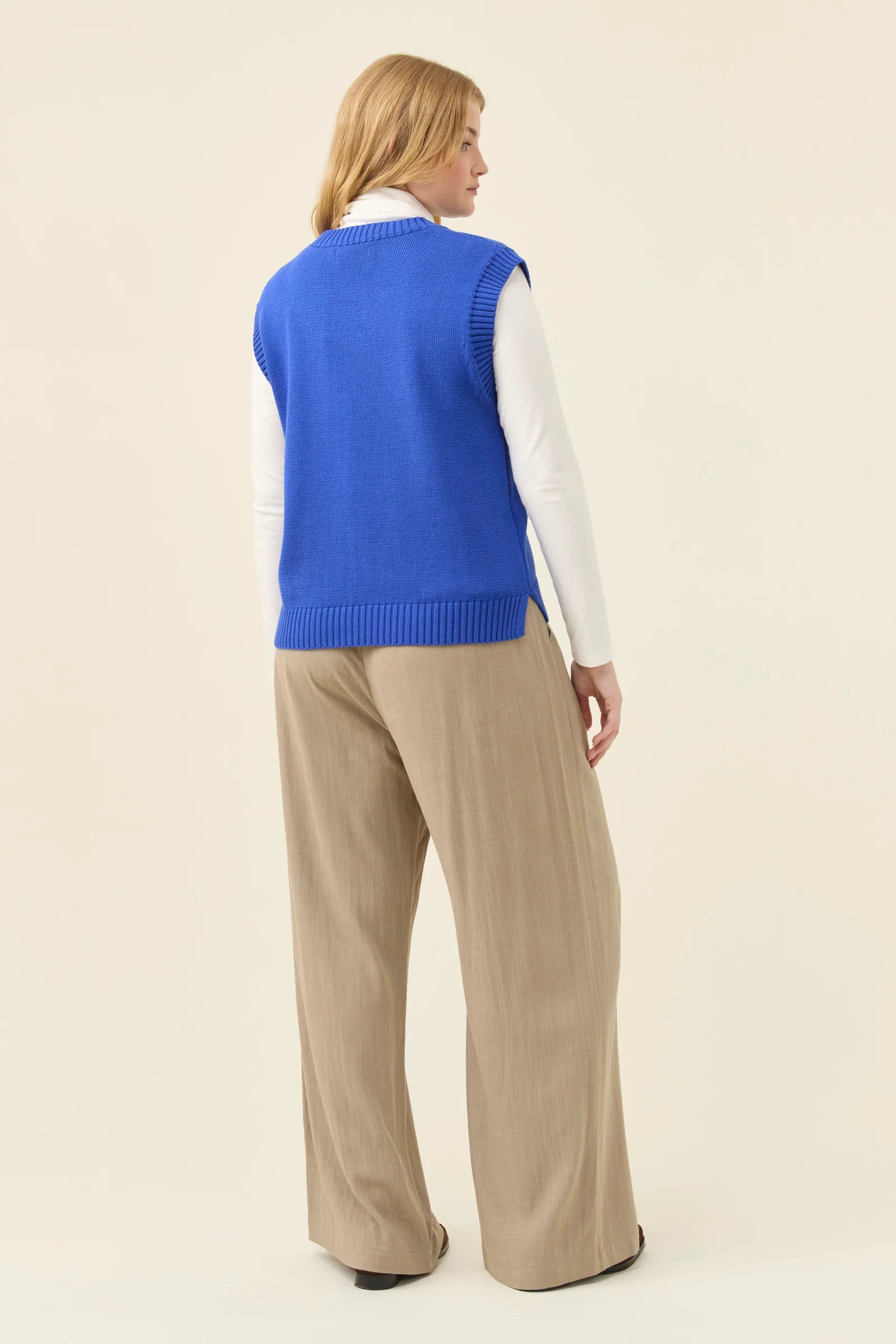 Woman wearing a Isle of Mine blue sweater vest over a white shirt and beige pants on a beige background
