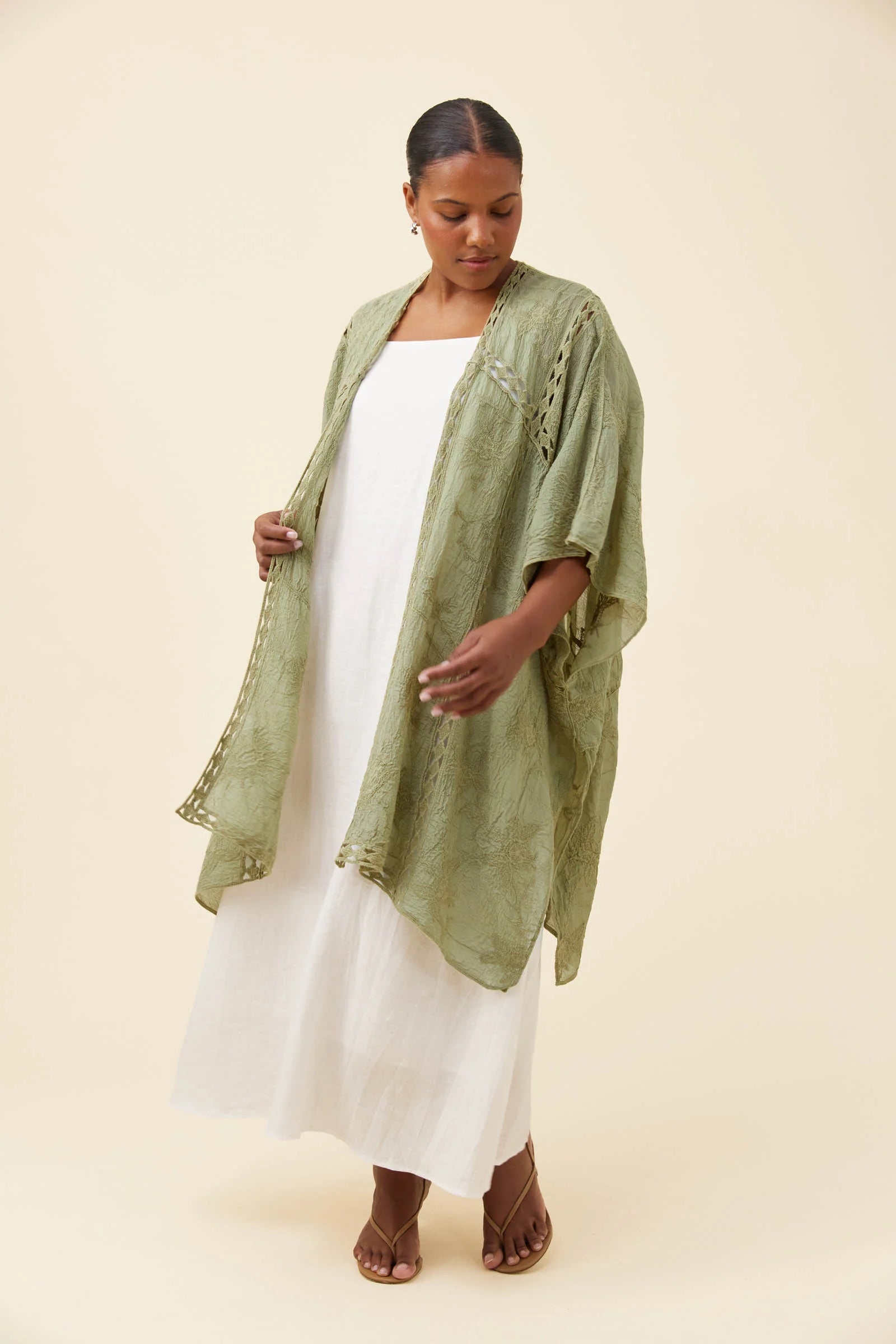 Image of A woman modeling a Isle of Mine sage green textured kimono cardigan over a white maxi dress, standing against a solid beige studio background.
