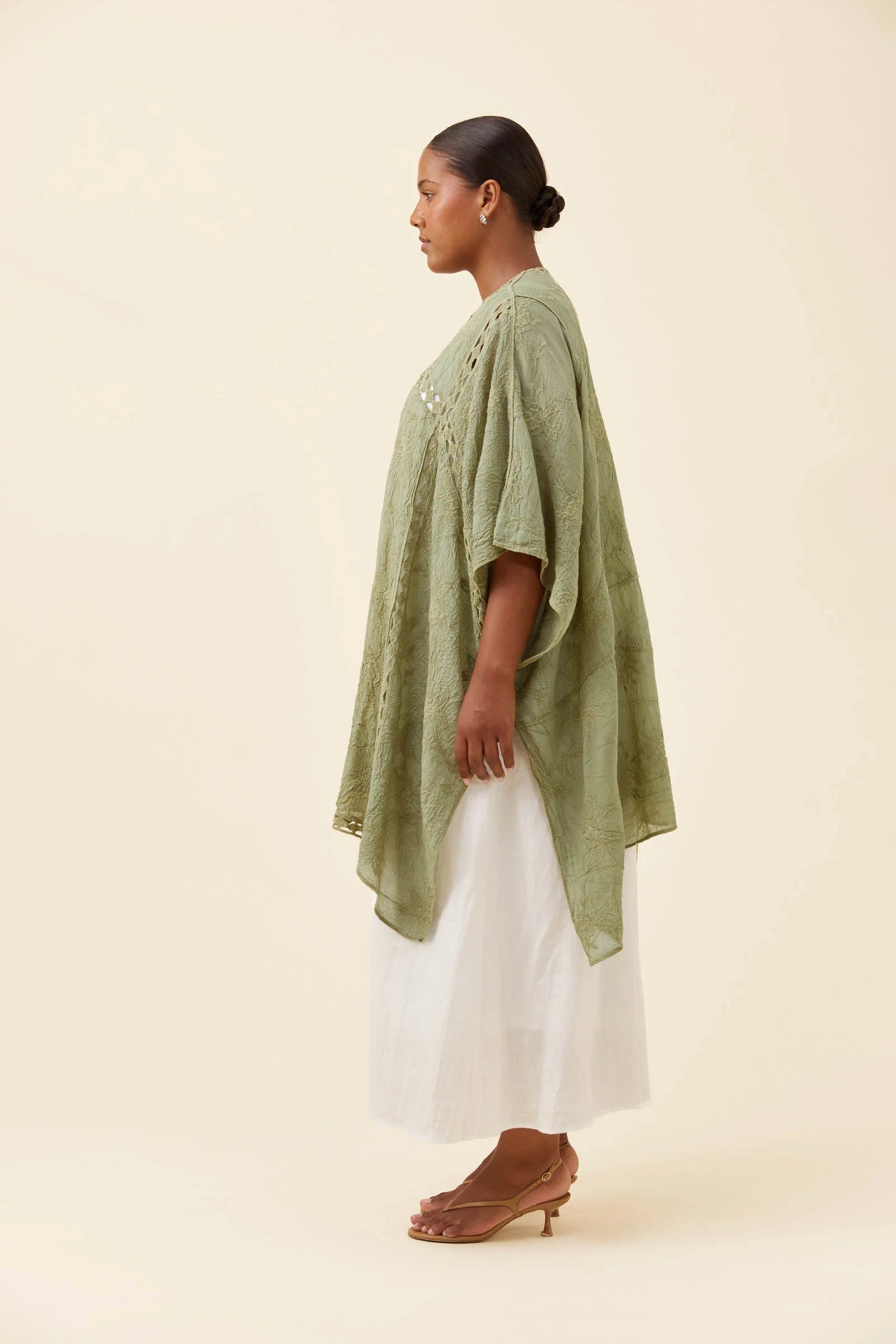 Image of A woman in profile wearing a Isle of Mine sage green embroidered tunic with a white skirt and brown heeled sandals, standing against a solid cream background.