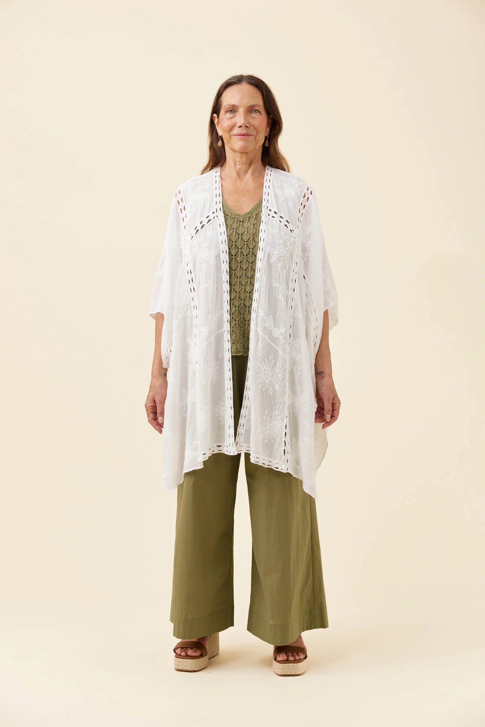 Image of A woman stands against a plain warm beige studio background wearing a Isle of Mine white open-front crochet or lace cardigan over an olive green top and matching olive green wide-leg pants.
