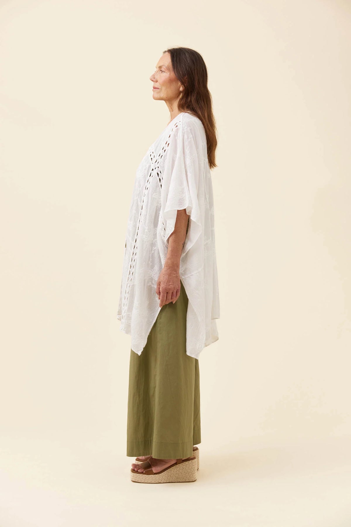 A woman stands in profile wearing a loose Isle of Mine white crochet-knit tunic over olive green wide-leg pants, with brown espadrille wedge sandals, against a plain warm beige studio background.