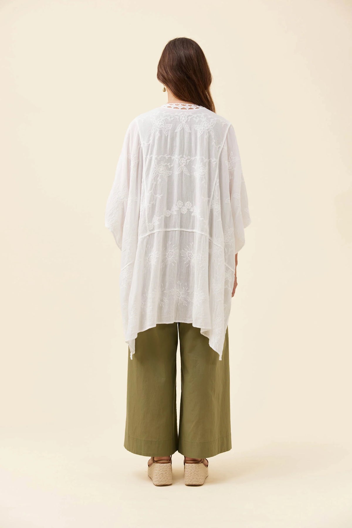 A woman viewed from the back wearing a Isle of Mine loose white embroidered tunic over olive green wide-leg pants and beige shoes, standing against a plain warm beige studio background.