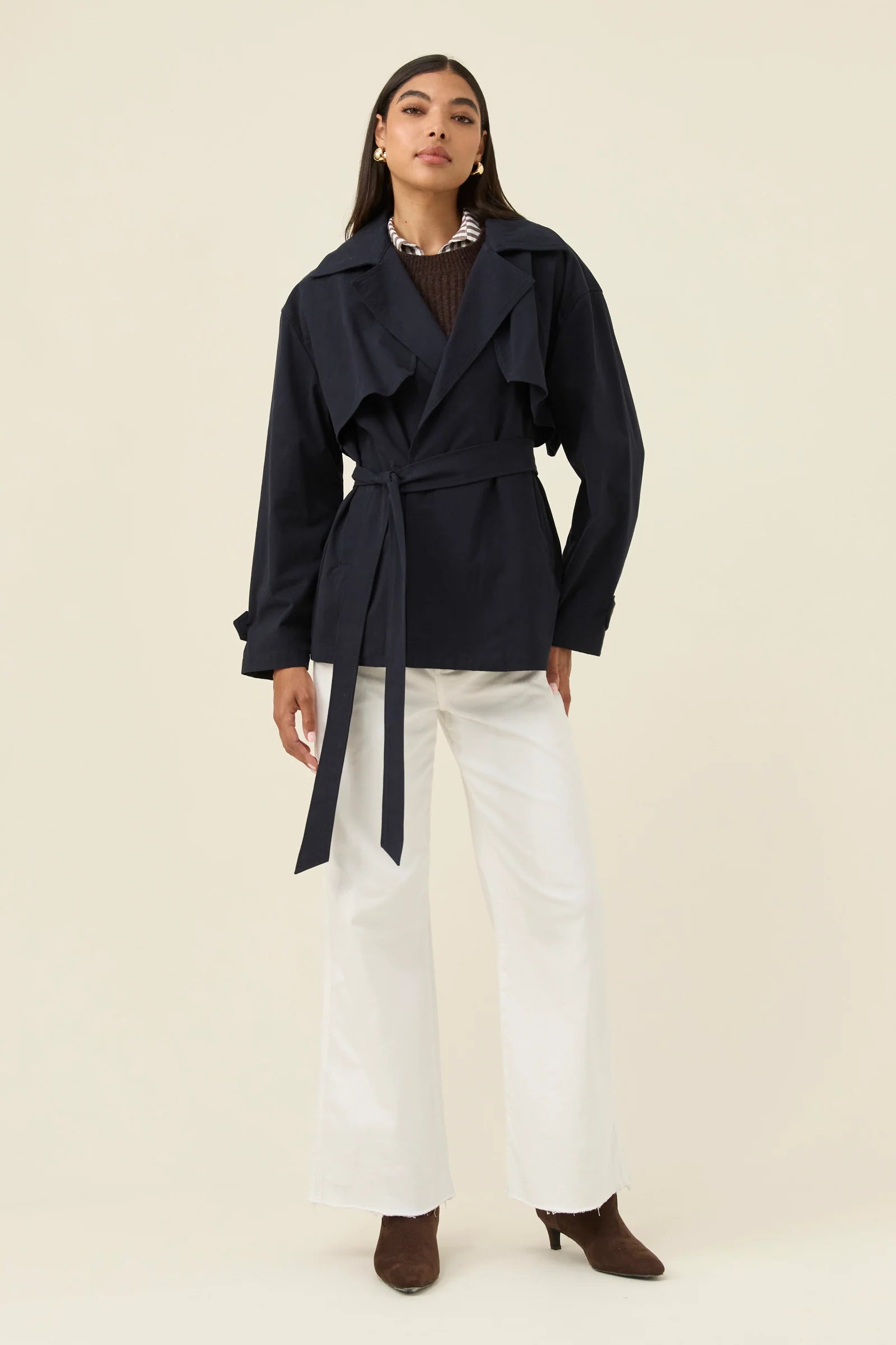 Woman wearing a Isle of Mine navy belted coat with white pants on a beige background