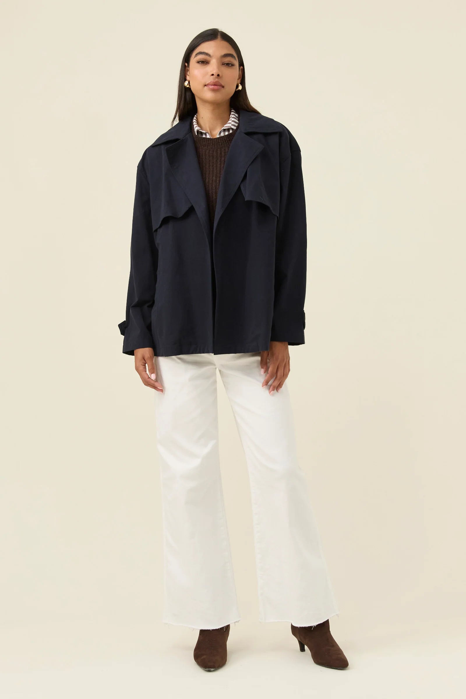 Woman wearing a Isle of Mine navy blue coat, brown sweater, white pants, and brown shoes on a beige background