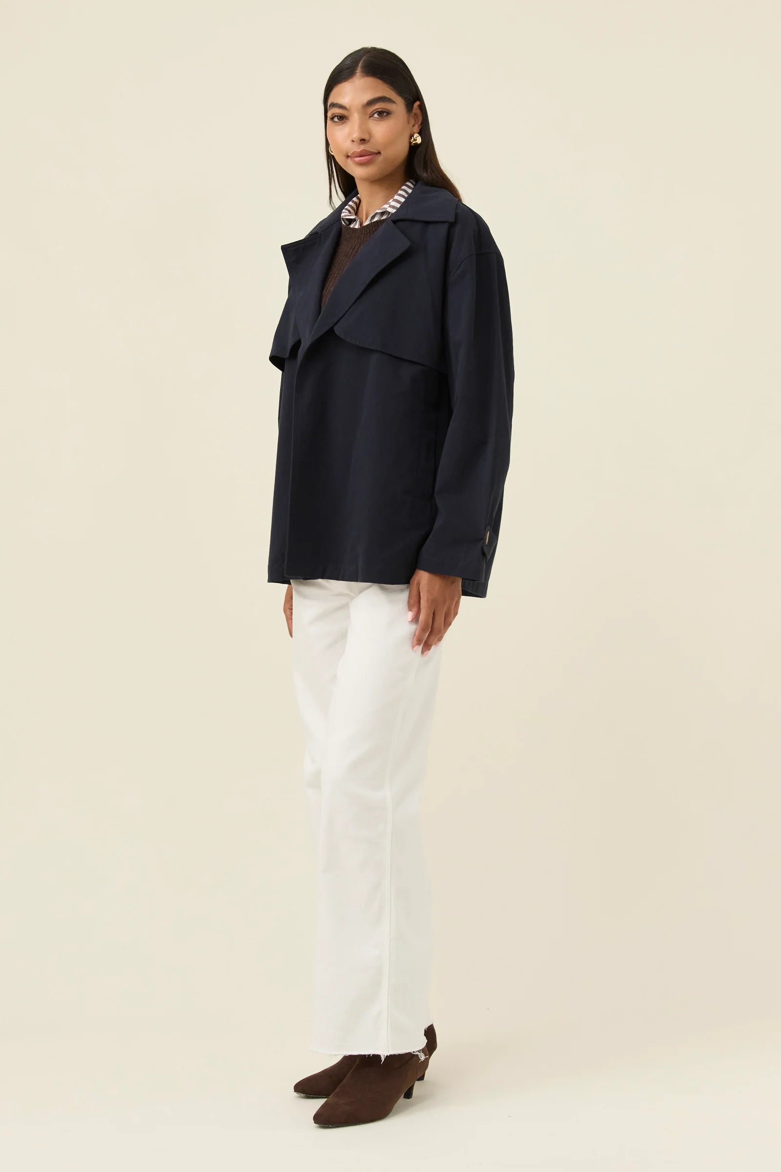Image of Woman wearing a Isle of Mine navy coat, white pants, and brown shoes on a beige background