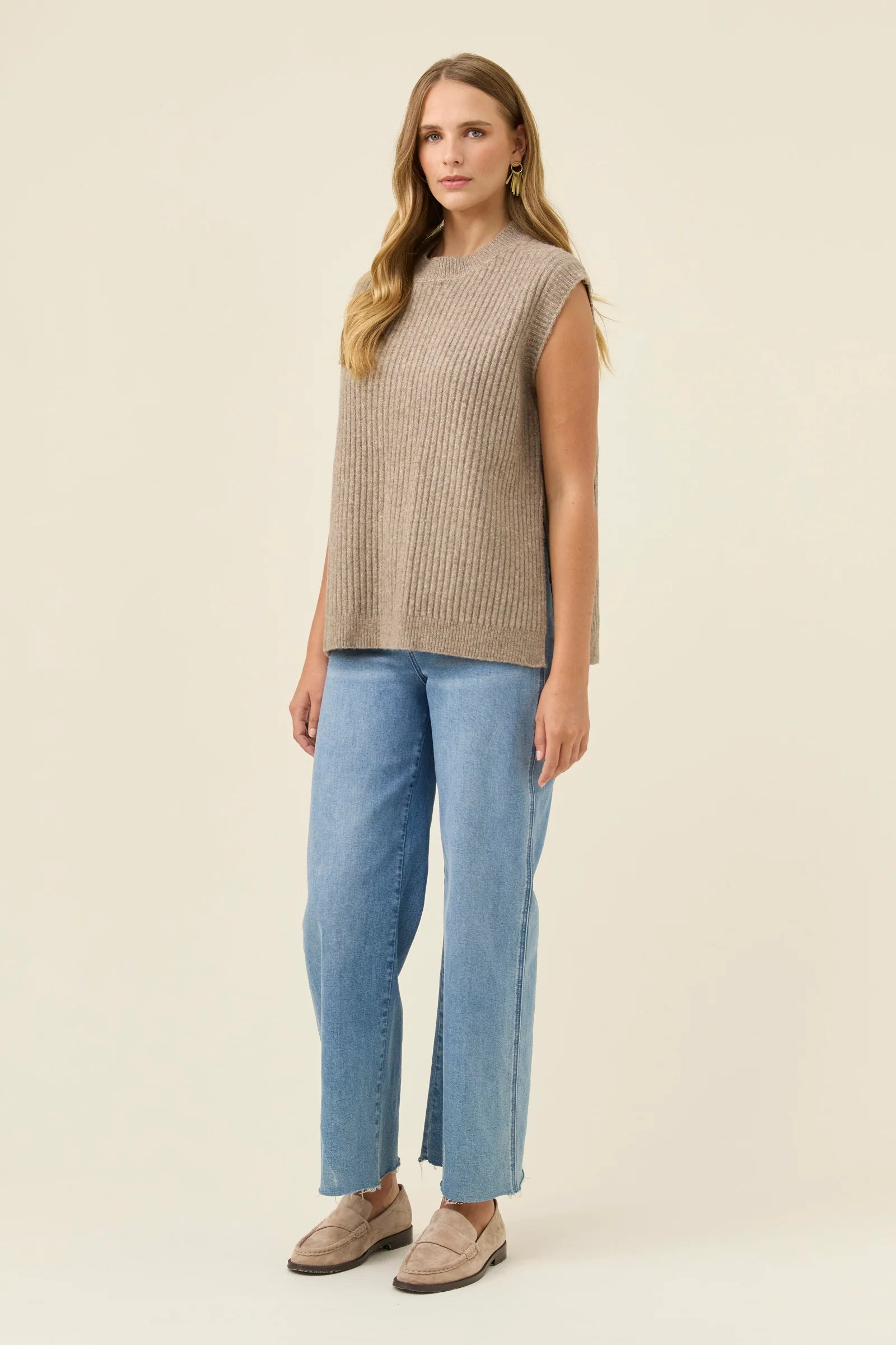 Woman wearing a Isle of Mine sleeveless beige knit top and blue jeans on a plain background