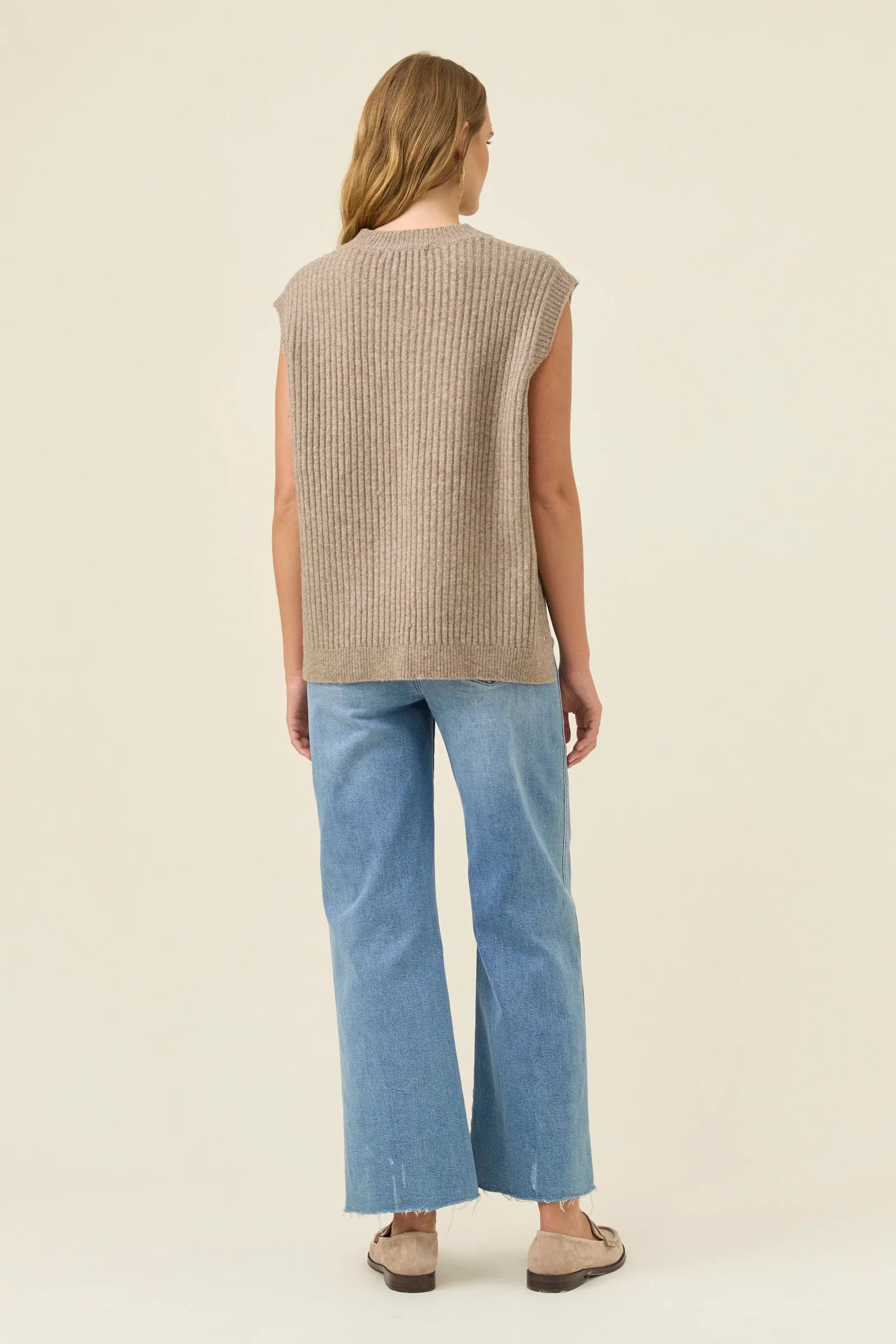 Woman wearing a Isle of Mine beige sleeveless sweater and blue jeans on a plain background