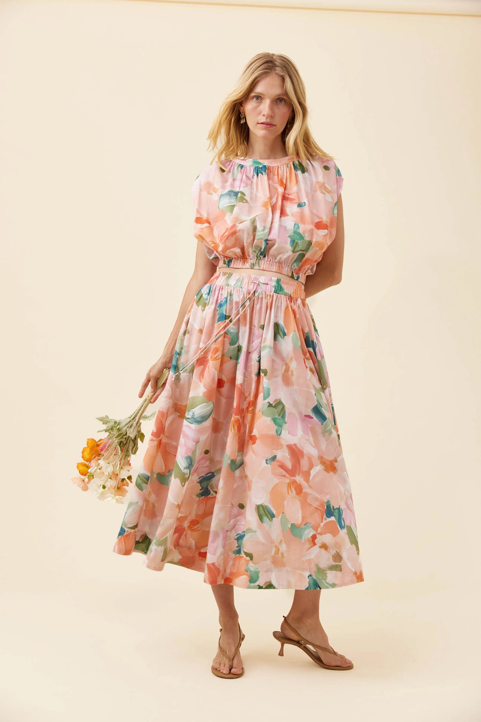 A woman modeling a Isle of Mine light pink skirt with a watercolor floral print in peach, orange, and green, holding a small bouquet of yellow and white flowers against a solid cream background.
