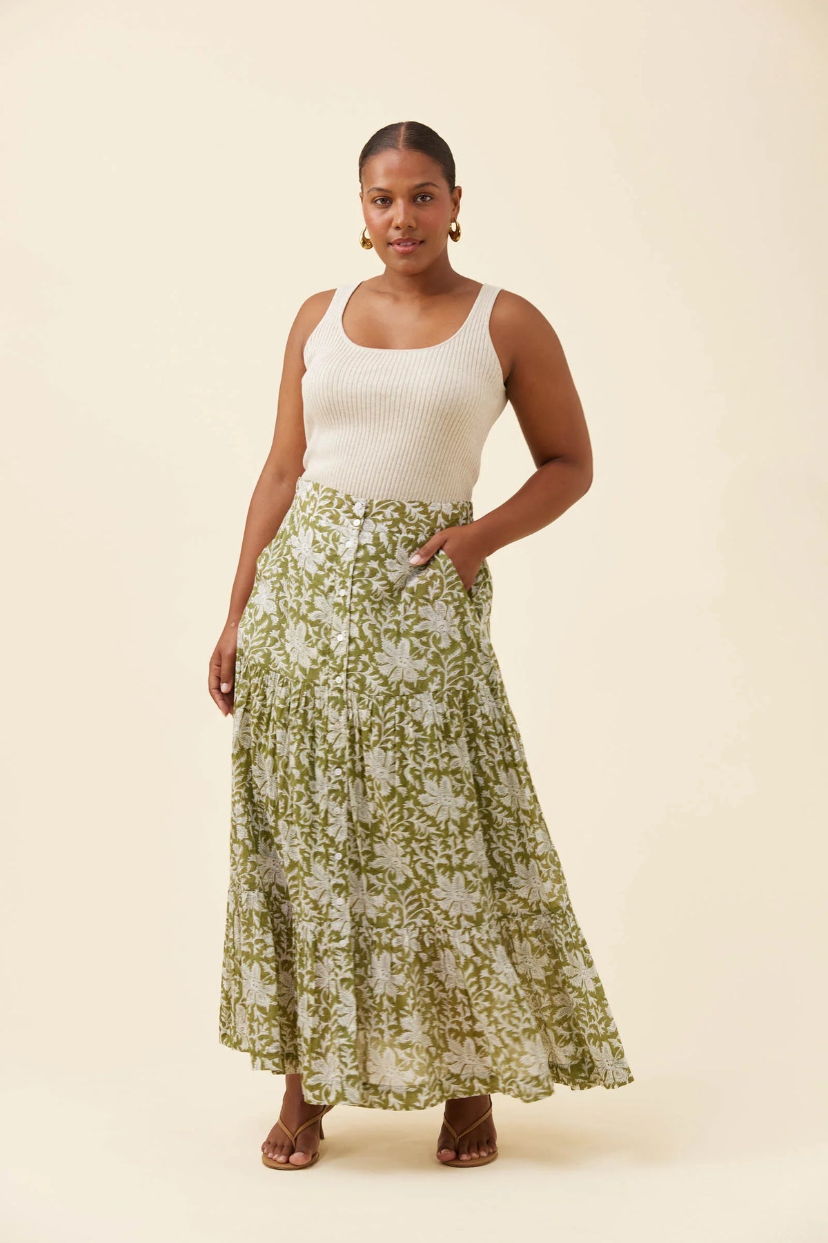 A woman modeling a white ribbed tank top and a Isle of Mine long, pleated maxi skirt with an olive green and white floral pattern, standing against a solid cream background.