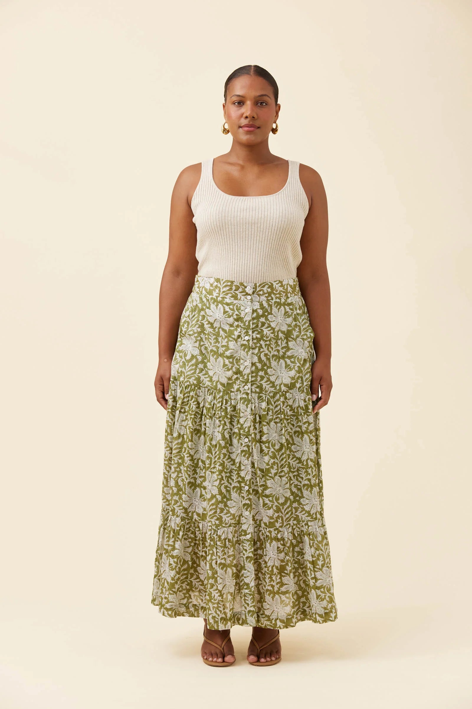 Image of A woman stands against a plain cream studio background wearing a sleeveless white ribbed tank top and a Isle of Mine long green floral maxi skirt with brown sandals.