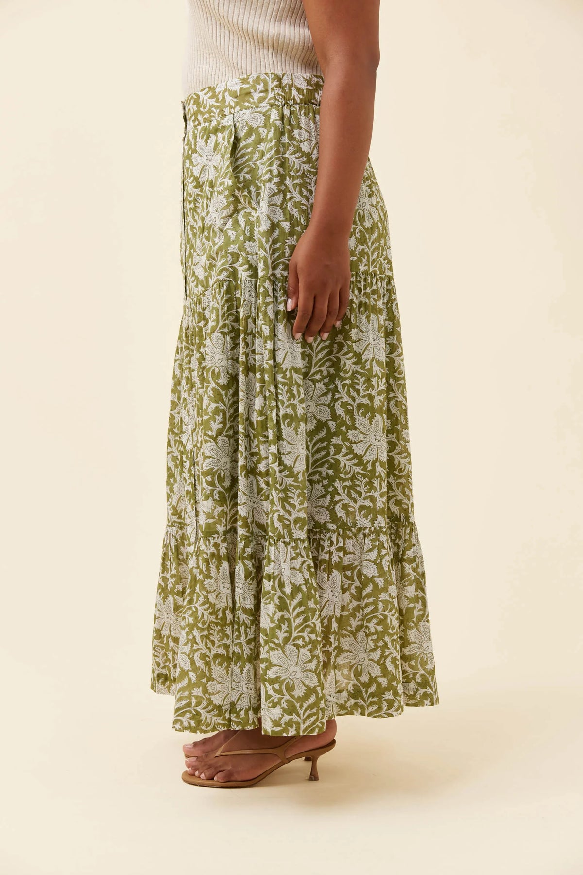 A model is shown from the waist down wearing a Isle of Mine long, tiered maxi skirt with an olive green and white floral pattern, paired with brown heeled sandals against a solid cream background.