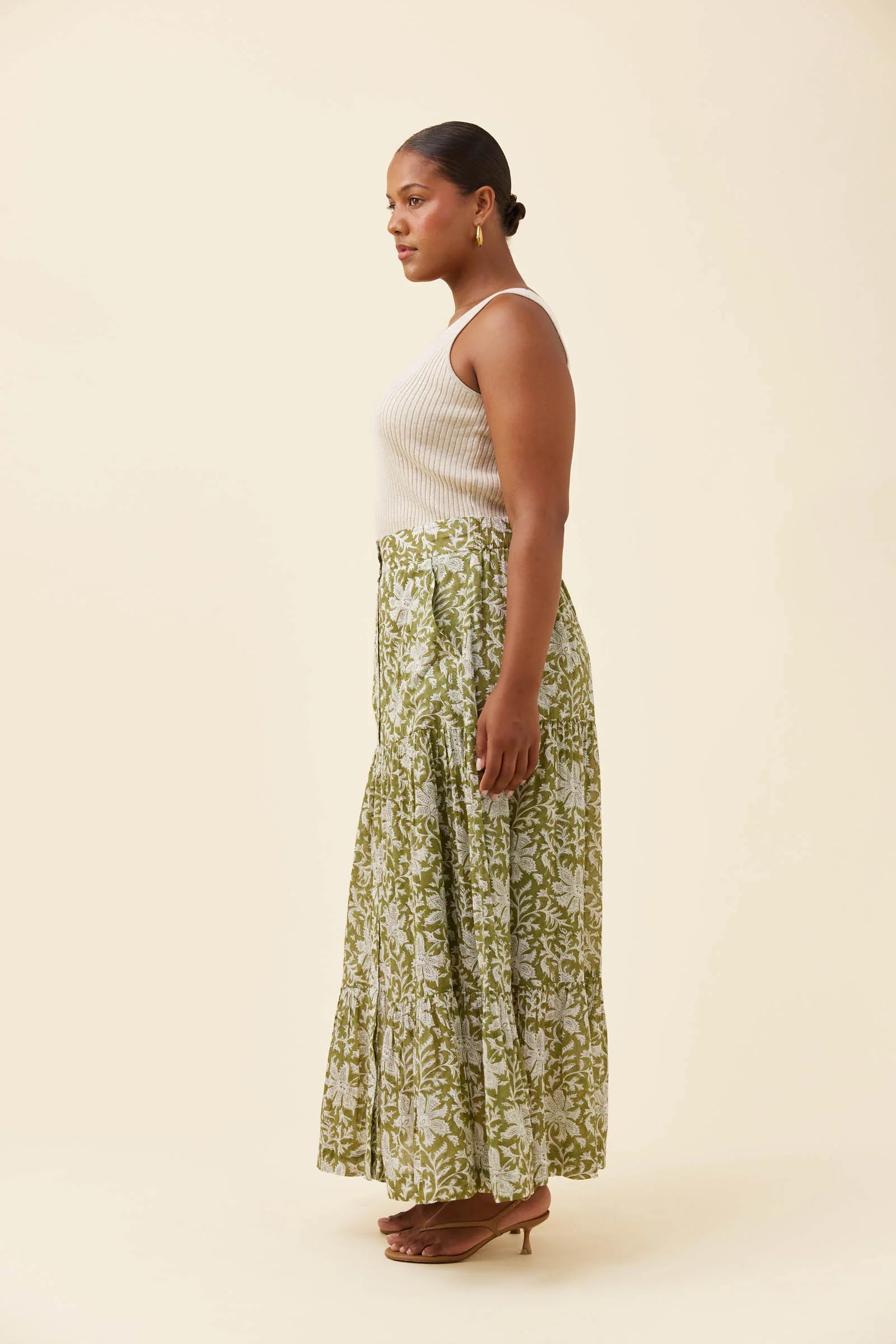 Image of A woman stands in profile wearing a sleeveless white ribbed knit top and a Isle of Mine long, flowing skirt with an olive green and white floral pattern. She is wearing brown heeled sandals against a solid, warm off-white studio background.
