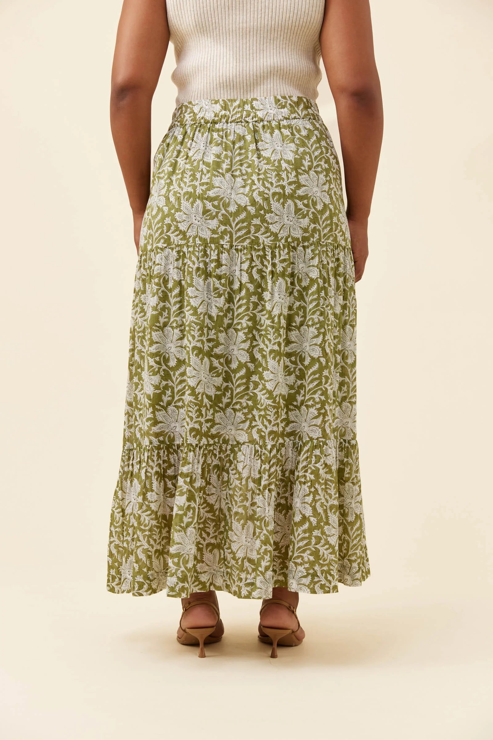 Image of A woman shown from the back wearing a Isle of Mine long, tiered maxi skirt with an olive green and white floral block-print pattern, paired with a cream ribbed top and brown heeled sandals, against a solid beige background.