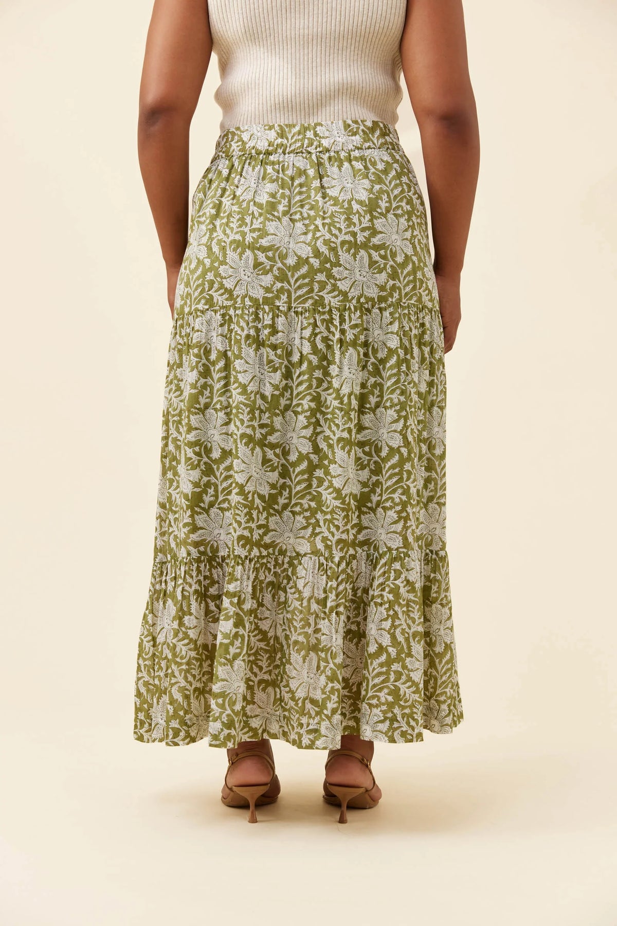 A woman shown from the back wearing a Isle of Mine long, tiered maxi skirt with an olive green and white floral block-print pattern, paired with a cream ribbed top and brown heeled sandals, against a solid beige background.