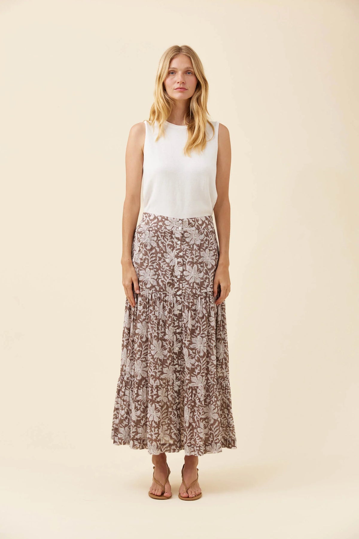 A woman stands against a plain beige studio background wearing a sleeveless white top and a Isle of Mine long pleated skirt with a brown and beige floral pattern.