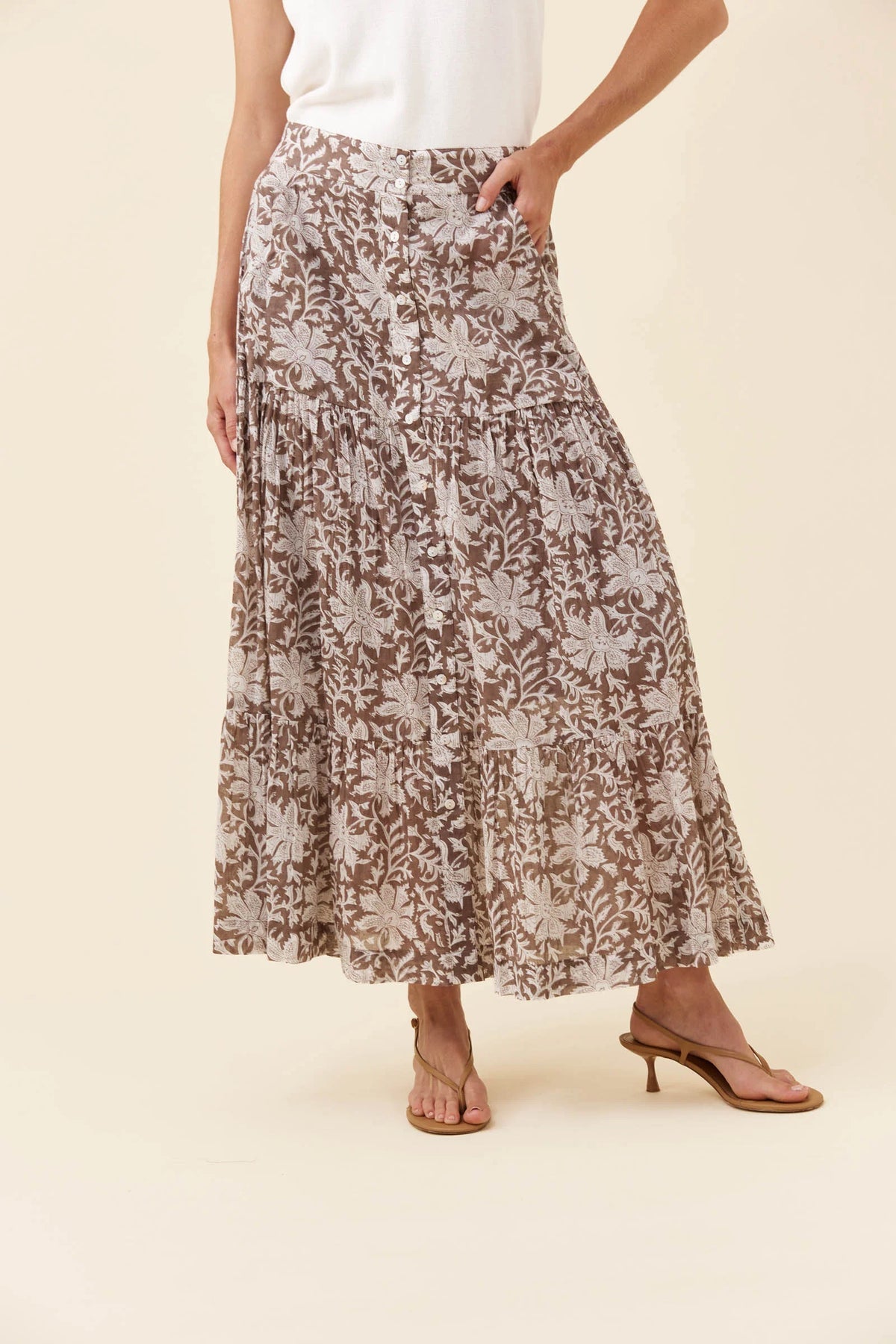 A woman wearing a Isle of Mine long, tiered maxi skirt with a brown and beige floral pattern, paired with a white top and brown heeled sandals, against a solid beige background.