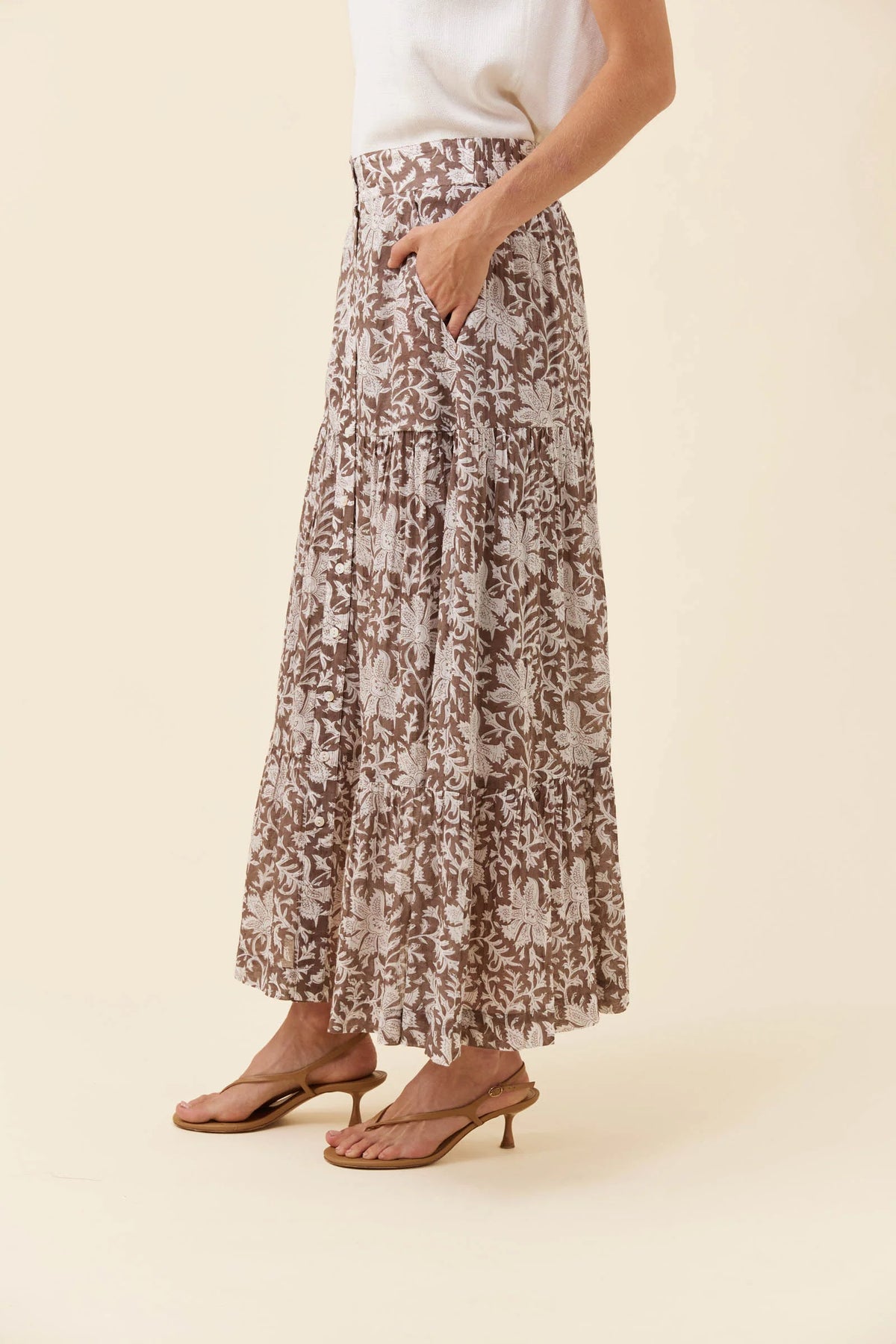 A model wearing a Isle of Mine brown and beige floral print maxi skirt with pockets, paired with a white top and brown heeled sandals, against a solid cream background.