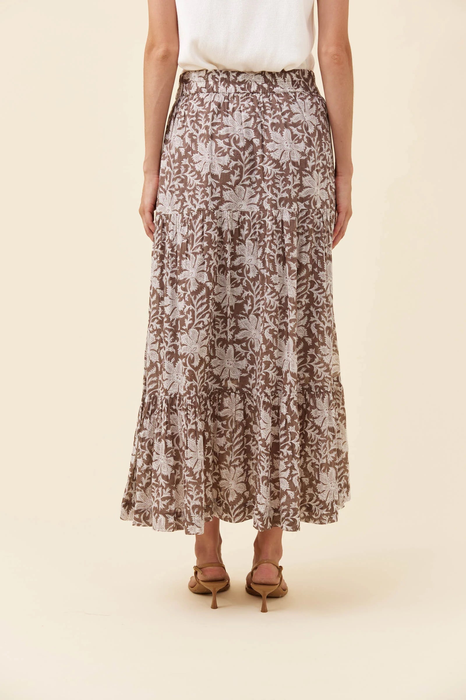 Image of A woman wearing a Isle of Mine brown and beige floral print maxi skirt with a white top and brown heeled sandals, shown from the waist down against a solid cream background.
