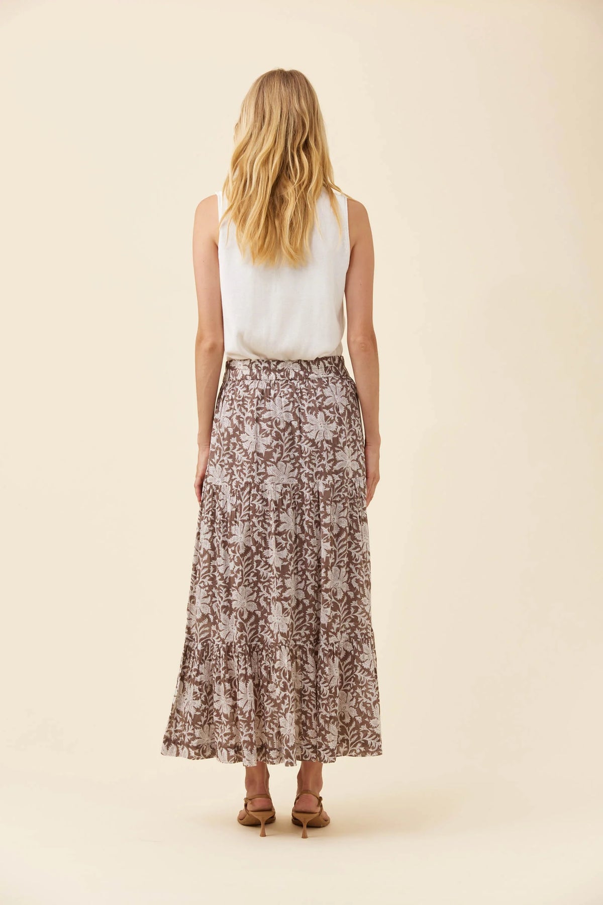 A woman shown from the back wearing a sleeveless white top and a Isle of Mine long brown floral print midi skirt, standing against a solid light beige background.