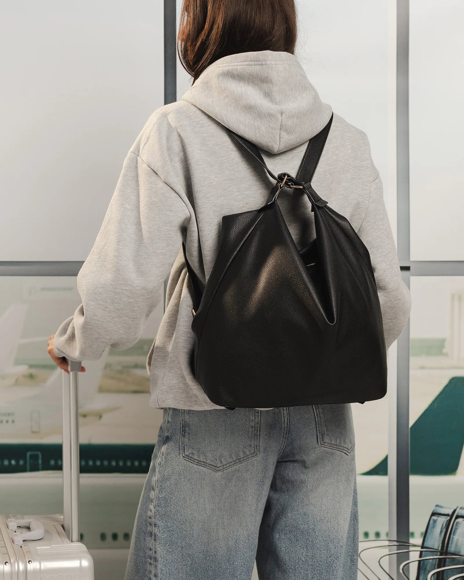 Woman wearing a Louenhide black drawstring bag and gray hoodie with a suitcase in an airport.