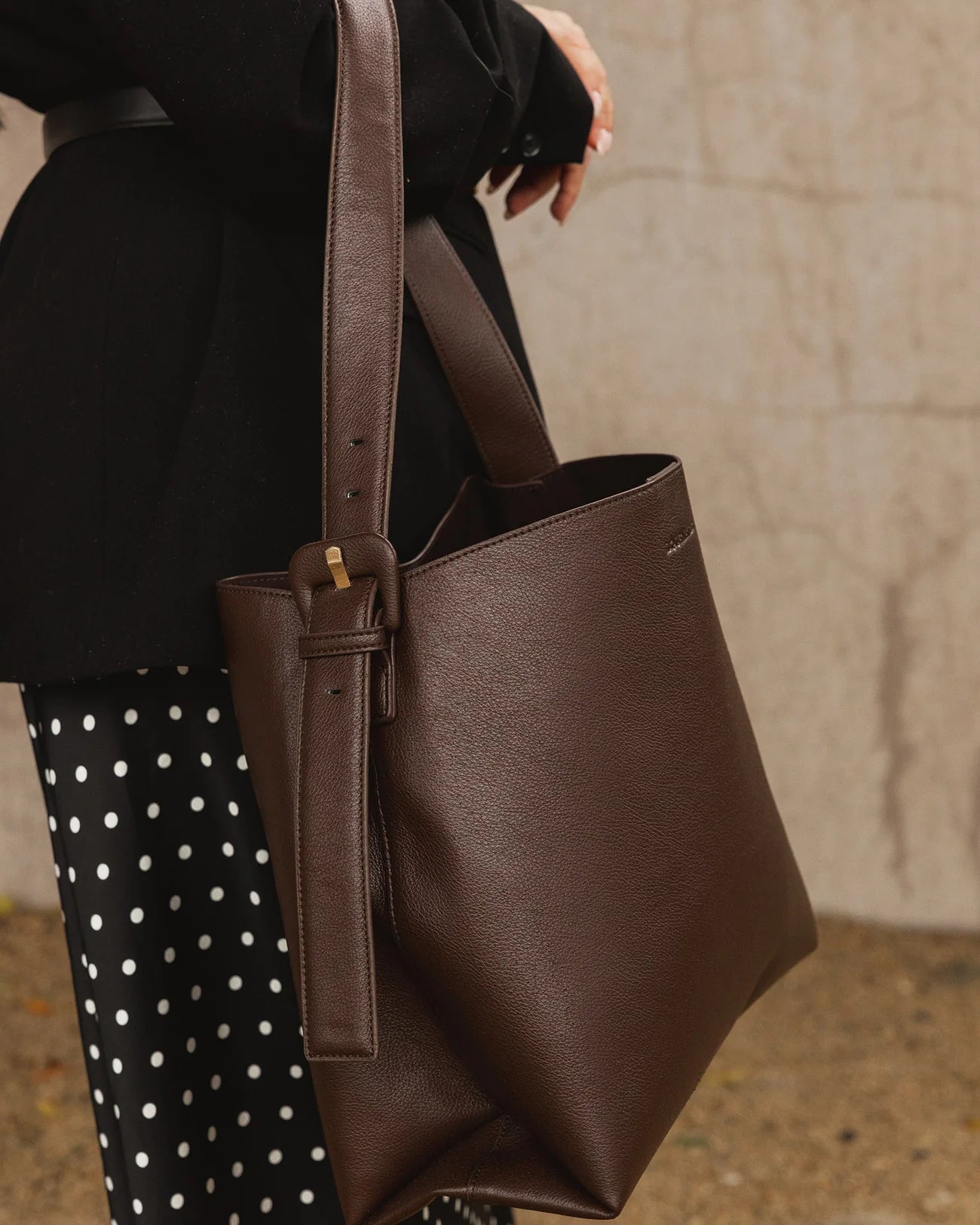 Louenhide brown leather tote bag held by a person wearing a black coat and polka dot pants.