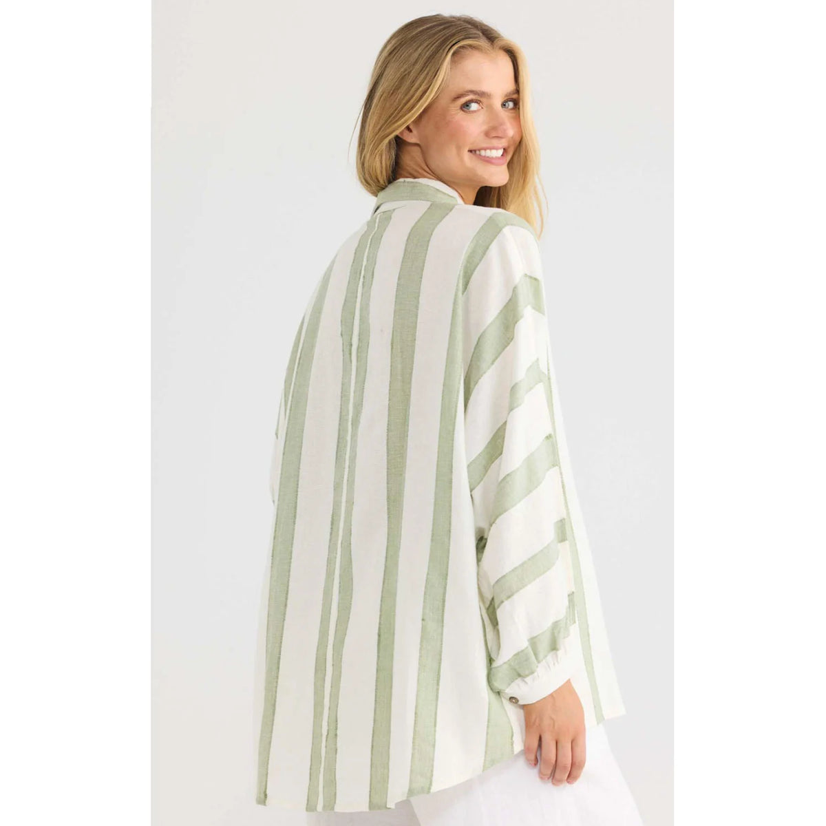 A woman wearing a Shanty Corporation loose, long-sleeve striped shirt in sage green and off-white, shown from a three-quarters back view against a plain light gray studio background.