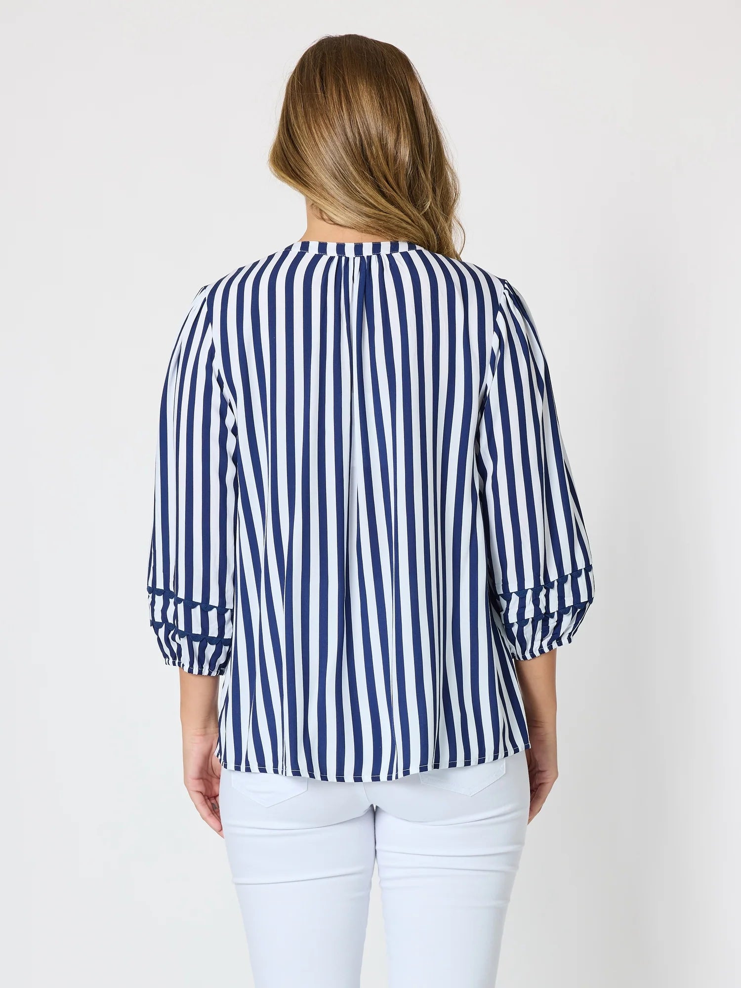 Image of A woman viewed from the back wearing a Threadz navy and white vertically striped blouse with three-quarter ruffled sleeves, paired with white pants, against a plain light gray background.
