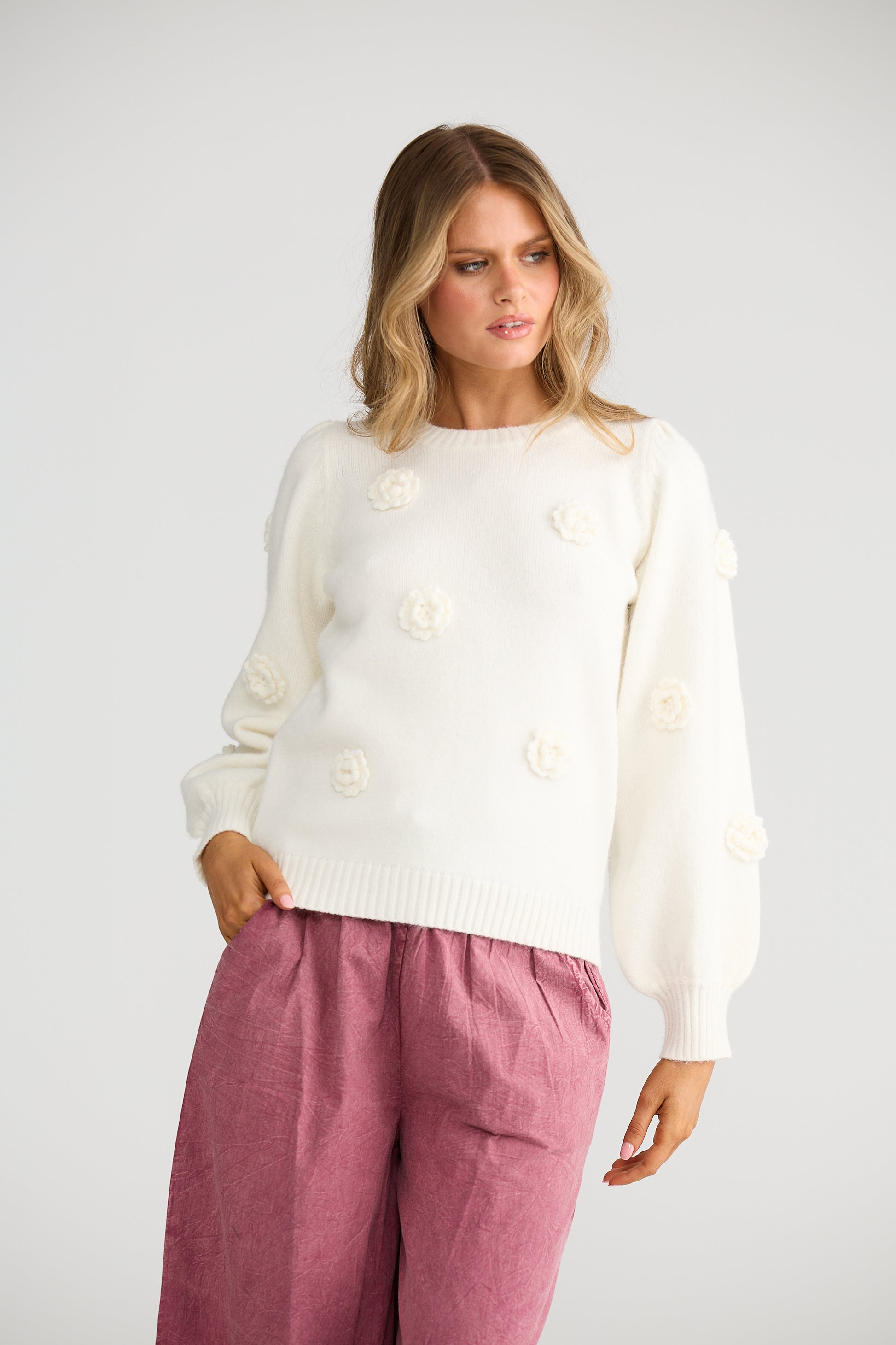 Woman wearing a Talisman white sweater with floral buttons and pink pants on a plain background